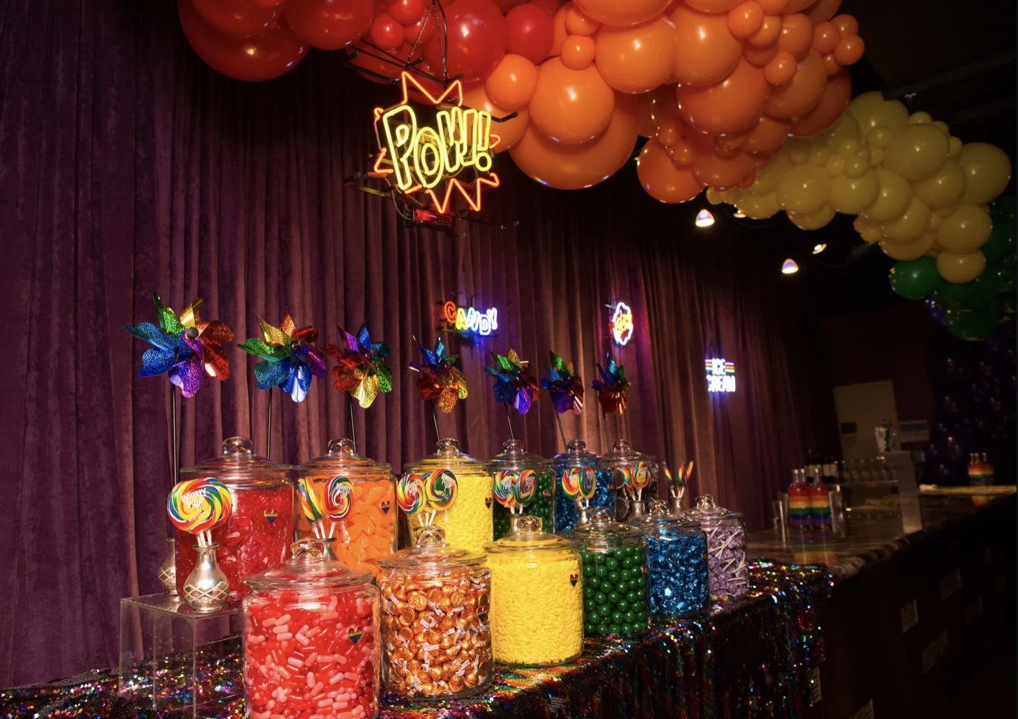 A colorful candy buffet with jars filled with assorted candies, lollipops, and rainbow-colored pinwheels on a sparkly tablecloth, with rainbow and themed neon signs and balloons overhead.