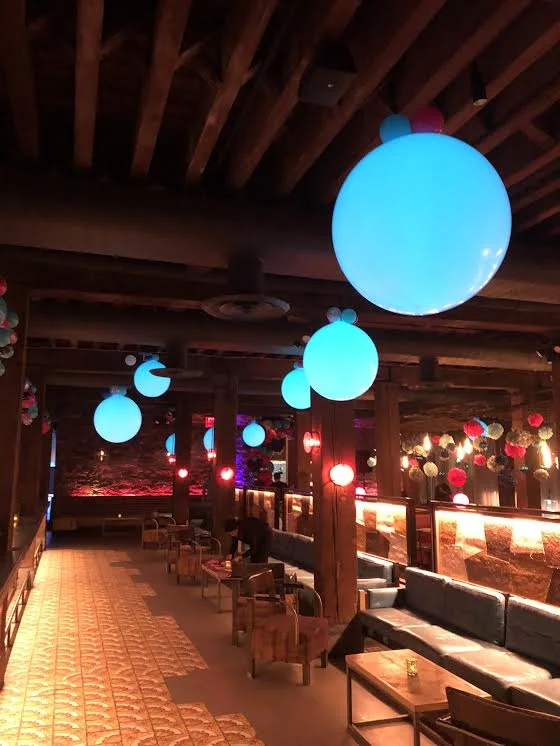 A dimly lit lounge with blue and red spherical hanging LED balloons, wooden beams on the ceiling, and a mix of sofas and chairs arranged along the walls.