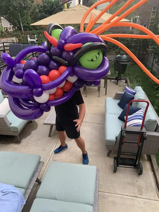 Person holding a large balloon sculpture of a colorful dragon with purple, green, red, black, and orange balloons in a backyard patio setting.