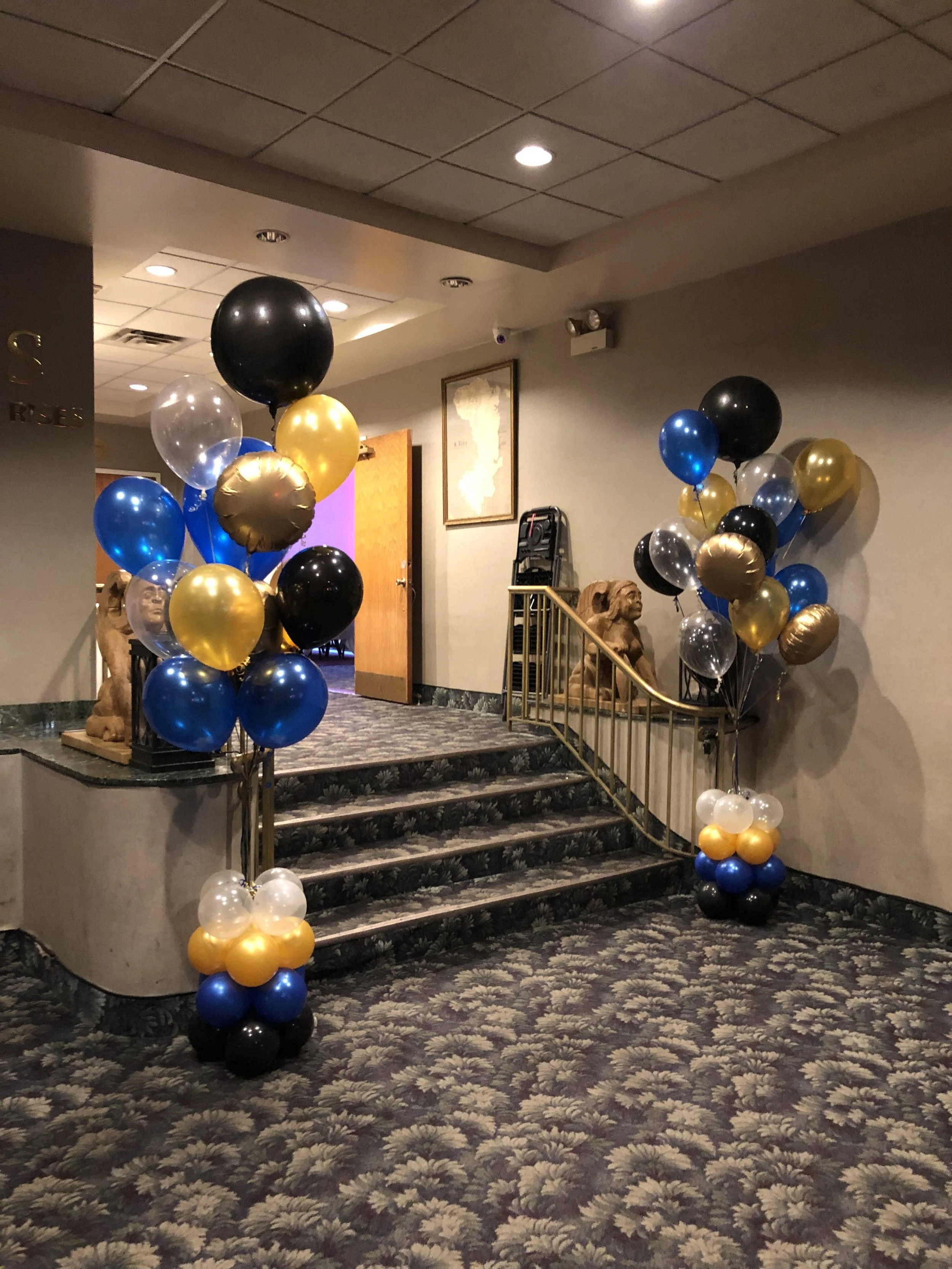 Decorated stairway entrance with gold, black, blue, and clear balloons in a banquet hall.