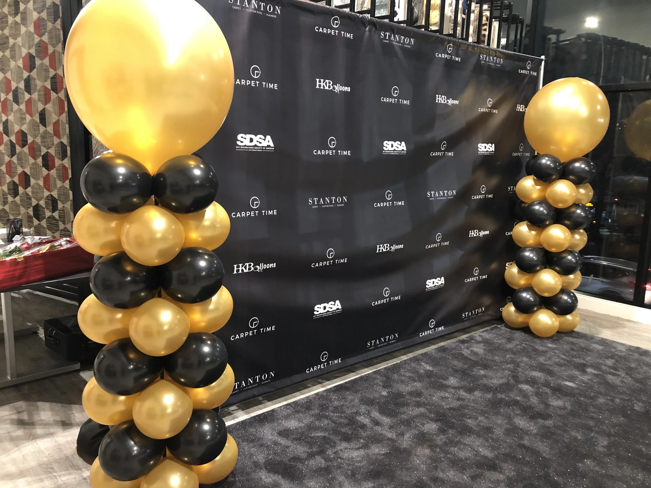 Black and gold balloon columns in front of a black backdrop with logos including Carpet Time, SDSA, Stanton, and HB Balloons for event decoration.