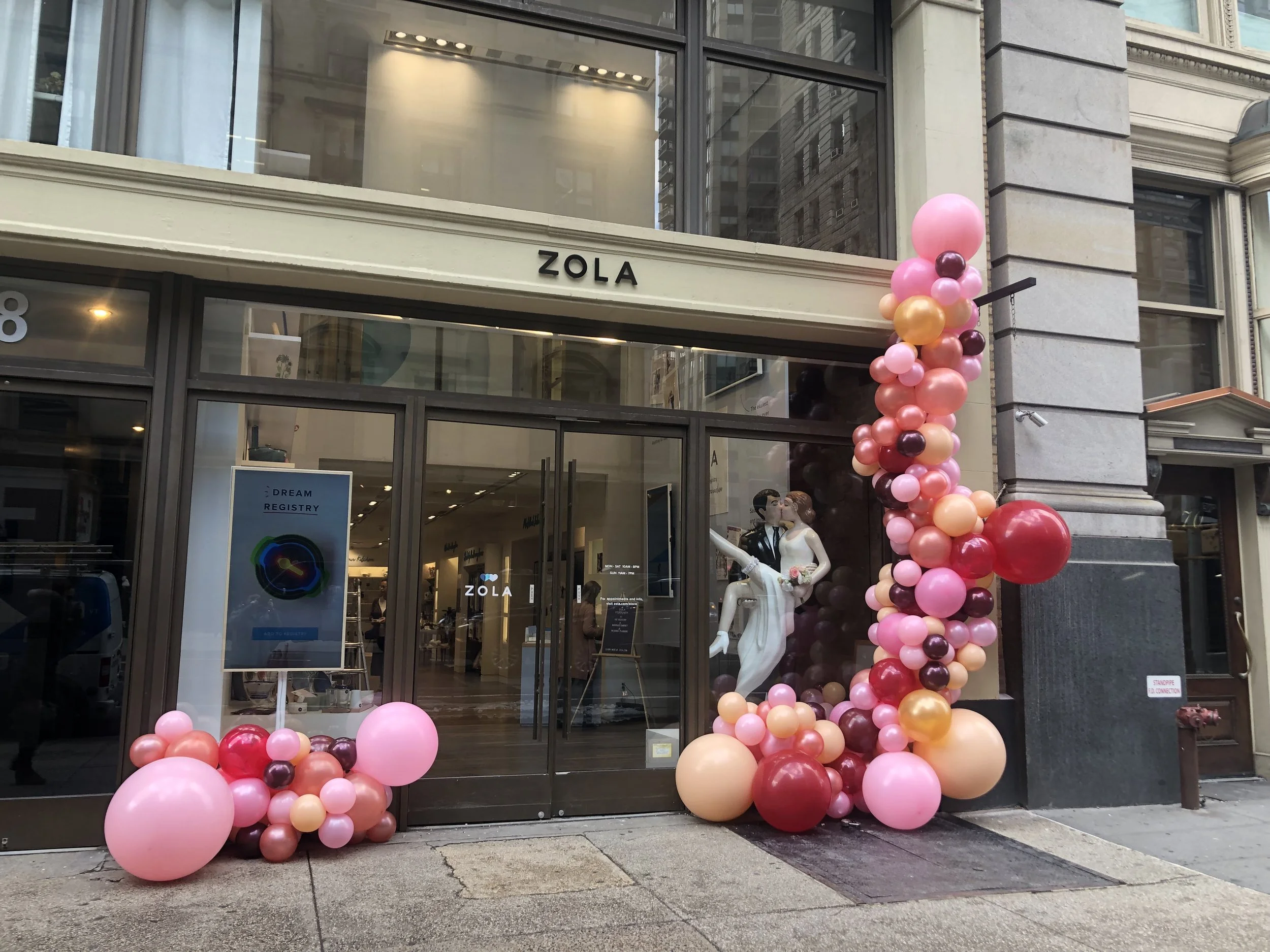 Storefront with pink, peach, burgundy, and red balloons arranged at the entrance. Mannequins dressed in wedding attire inside, with the store name ZOLA above the glass doors.
