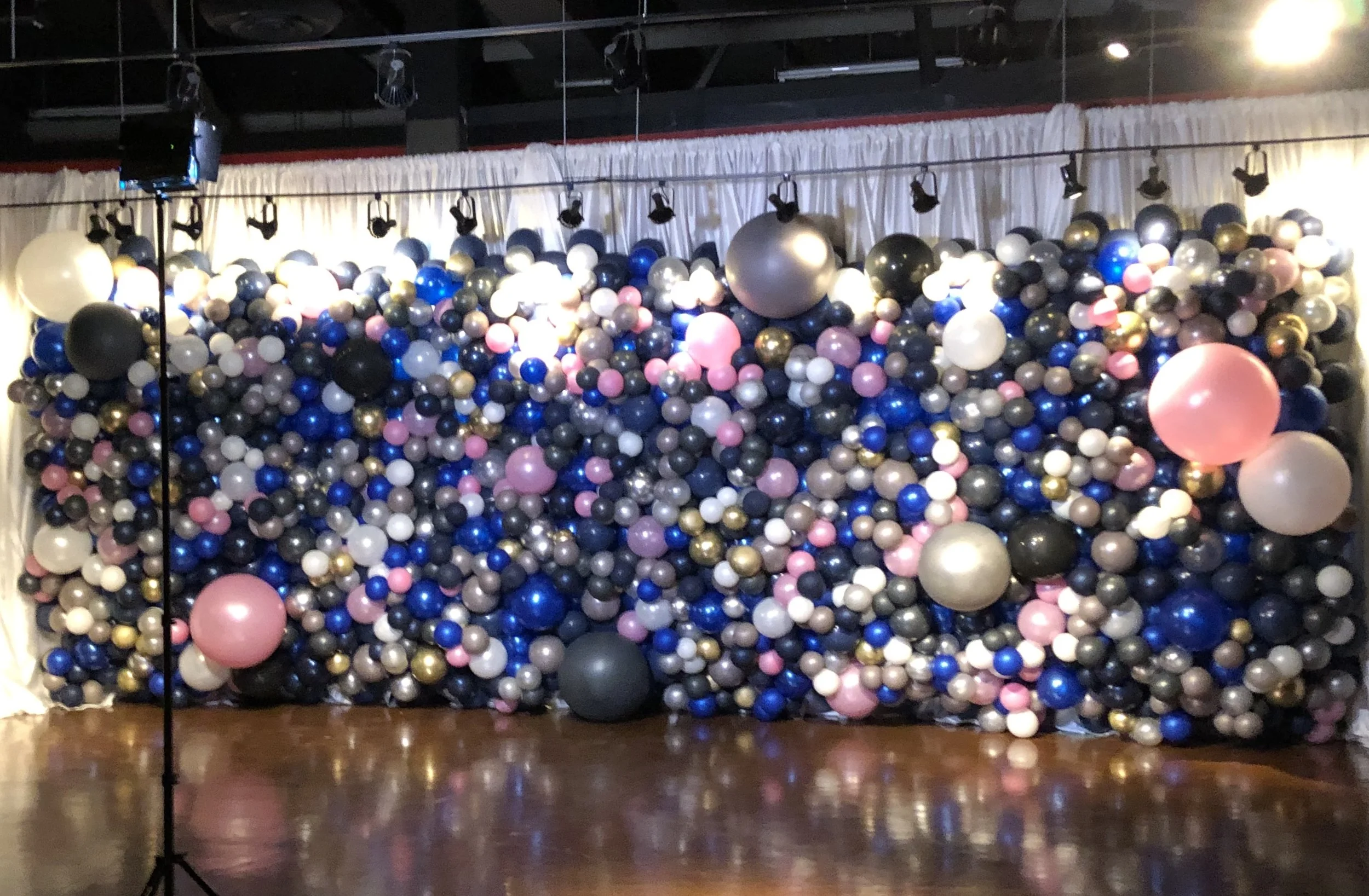 A wall decorated with numerous balloons in various colors including black, white, pink, blue, gray, and gold, with some balloons larger than others, arranged behind a stage with a white curtain and overhead lighting.