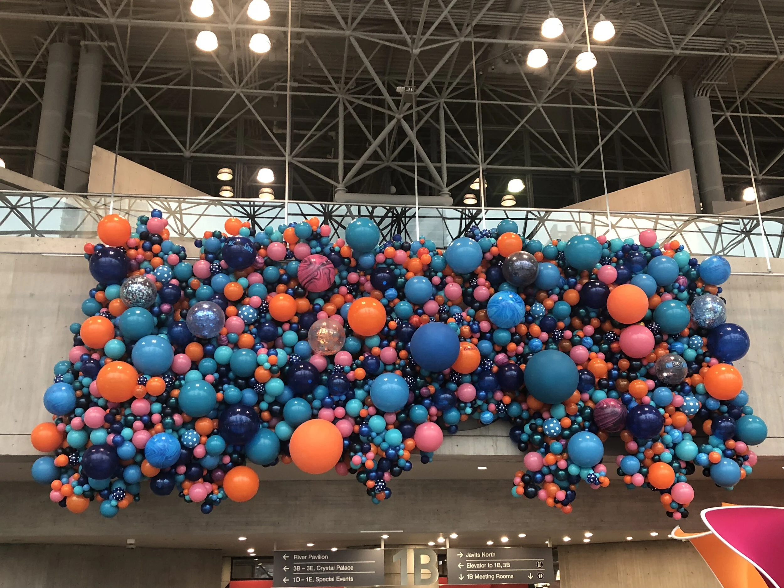 Colorful balloon art installation featuring numerous balloons in shades of blue, pink, orange, purple, and silver, arranged in a clustered display on an indoor wall with a modern ceiling and lighting structure in the background.