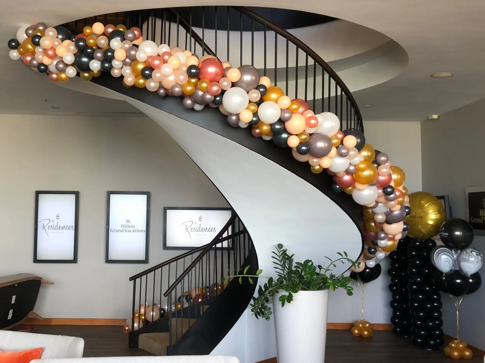 Balloon decorations including a cluster of multicolored balloons on a curved staircase railing, with more balloons on the floor and against the wall, in a decorated room with framed signs and a potted plant.