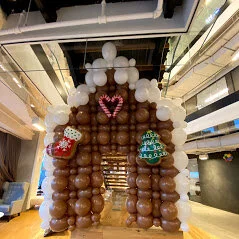 Balloon arch decorated with white, brown, and heart-shaped balloons creating a gingerbread house shape, with candy and stocking balloons attached.