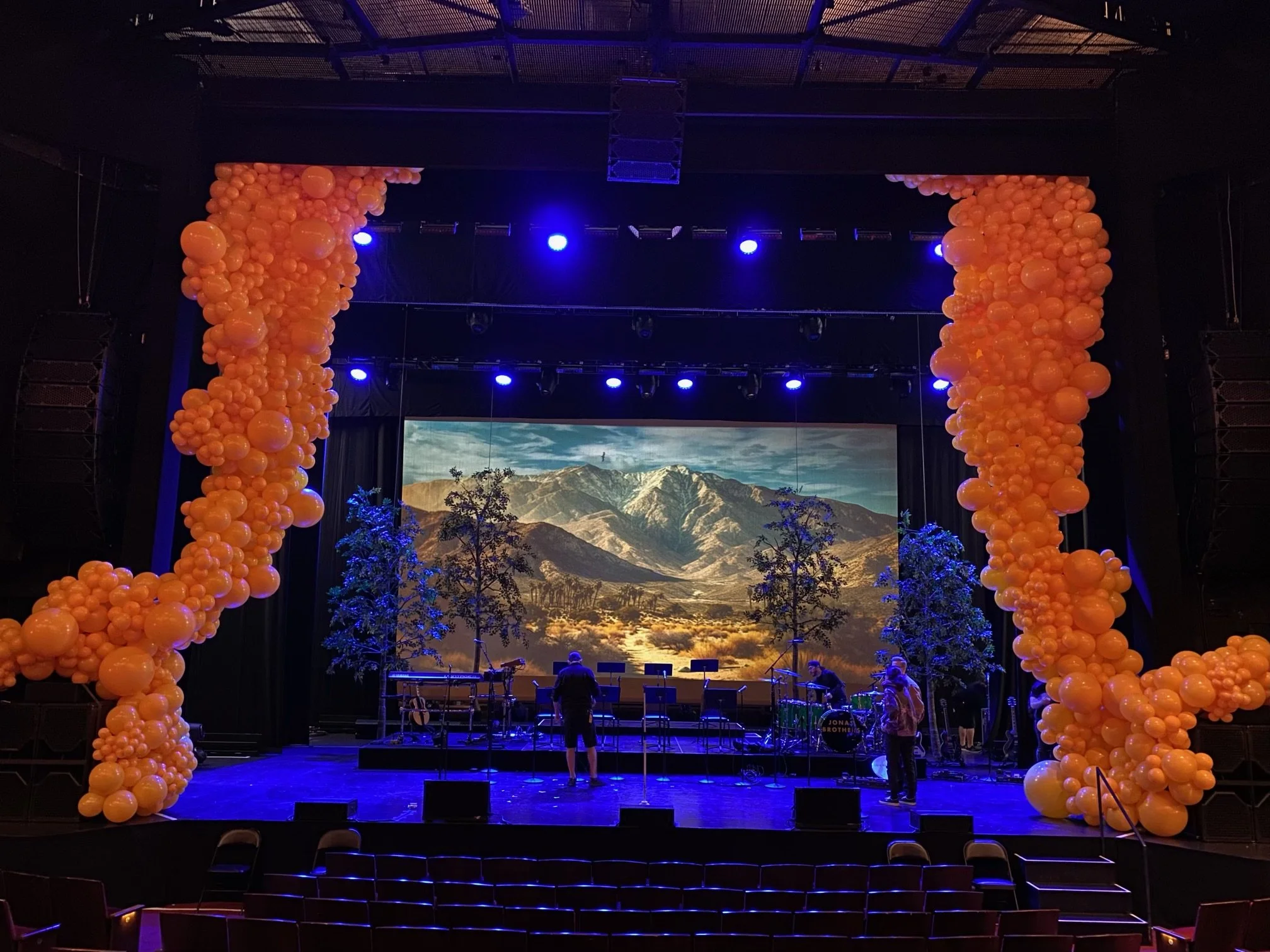 Stage decorated with large orange balloon arrangements on sides, with a backdrop of mountains and a sky projected on a screen. There are trees and musical instruments on the stage, and people are preparing for a Jonas Brothers performance.