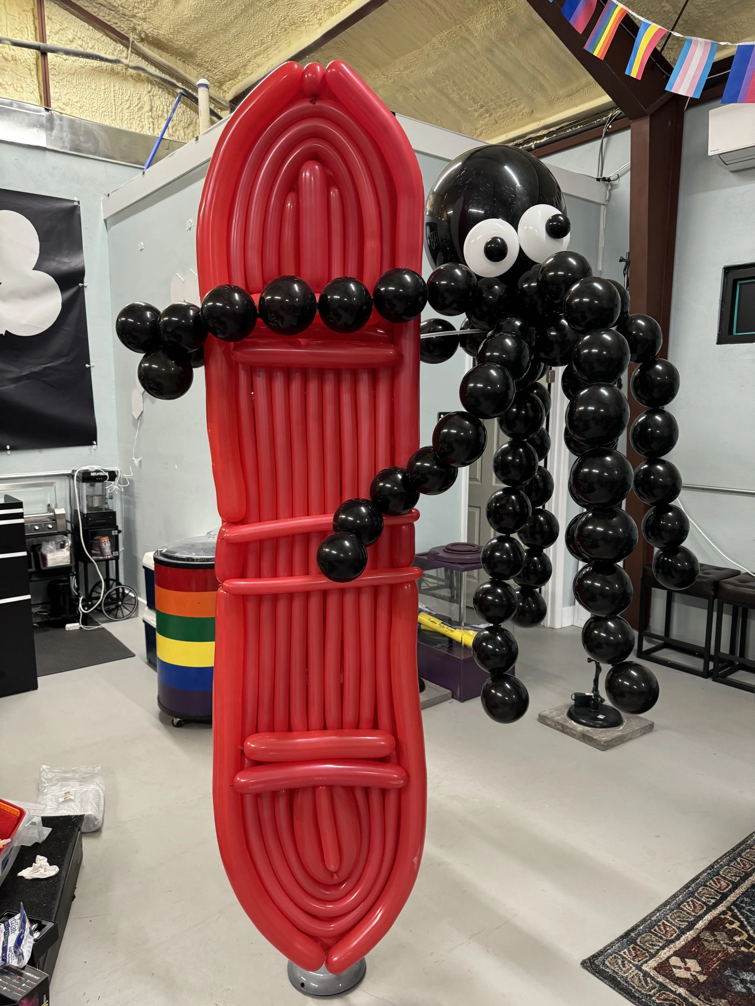 A large balloon sculpture of a black octopus holding a red surfboard, made of black, white, and red balloons, is displayed indoors against a yellow ceiling and gray walls.