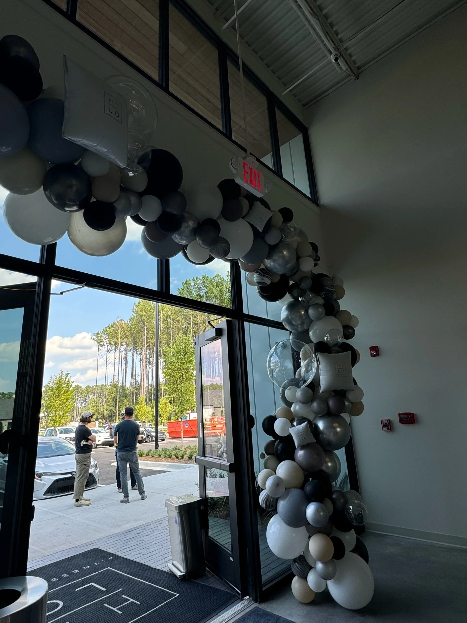 Balloon arch and decorations near the entrance of a building with large glass doors and windows.