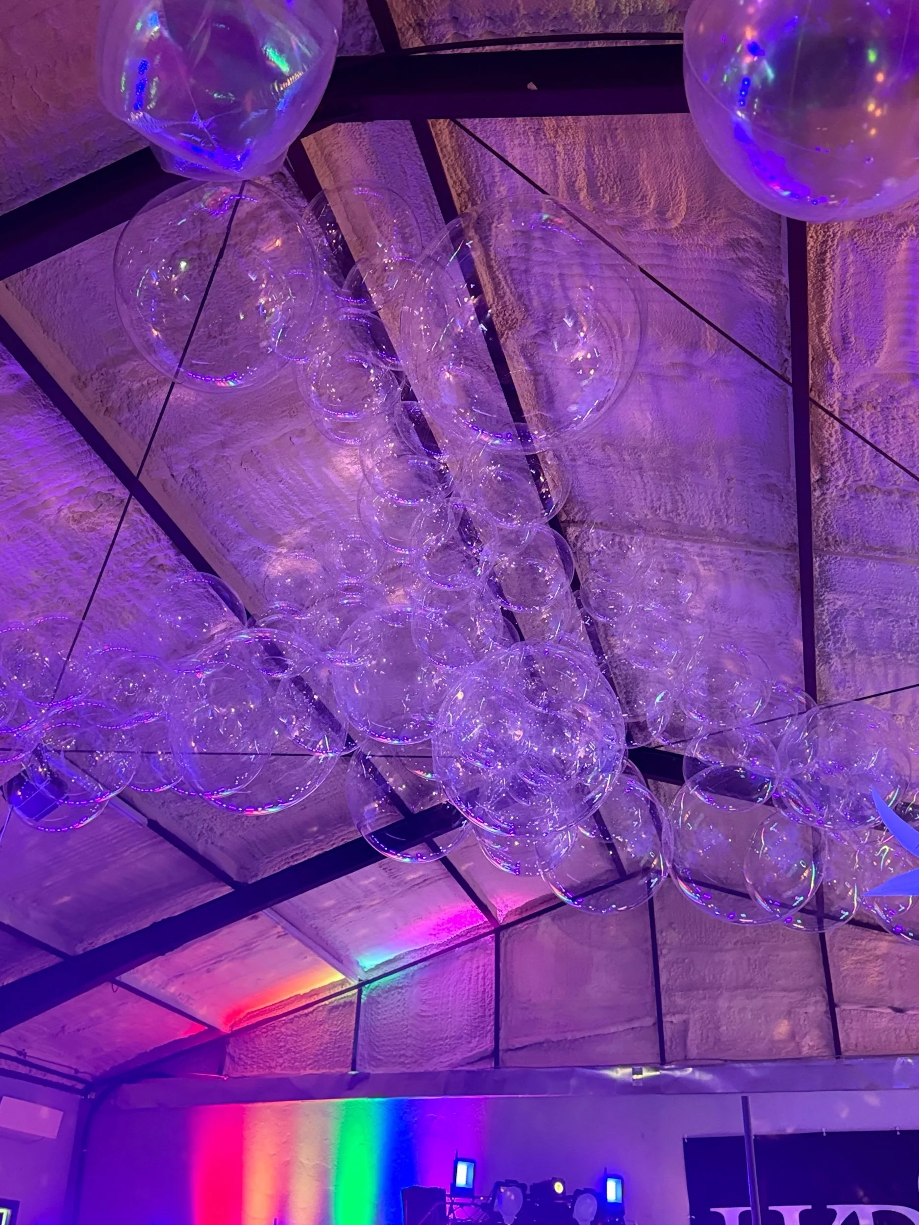 A ceiling decorated with a chandelier of multiple transparent balloons illuminated with purple and multicolored lights.