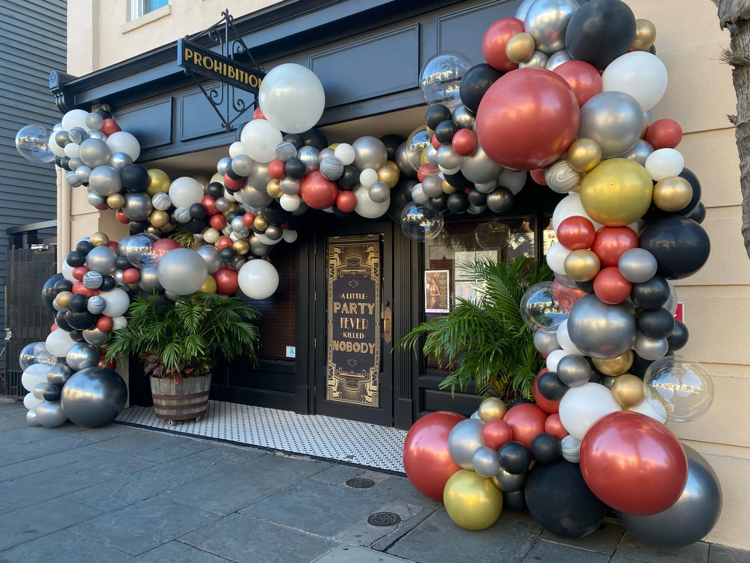 Entrance decorated with an arch of balloons in black, gold, white, red, and clear, with potted plants on either side of the door and a sign that reads 'A Little Party Never Killed Nobody'.
