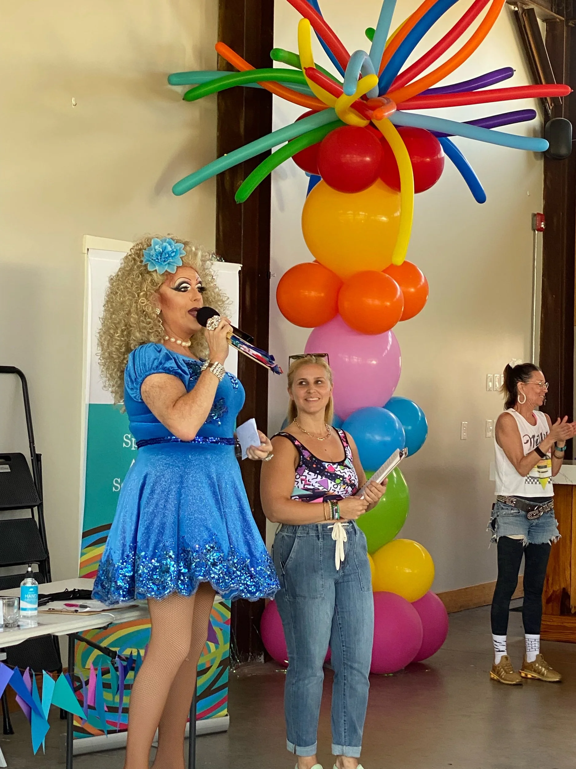 A person dressed as a drag queen with curly blond hair, blue dress, and accessories holding a microphone, standing next to a woman with blonde hair, glasses, and casual clothing, in front of a colorful balloon sculpture in a decorated indoor space.