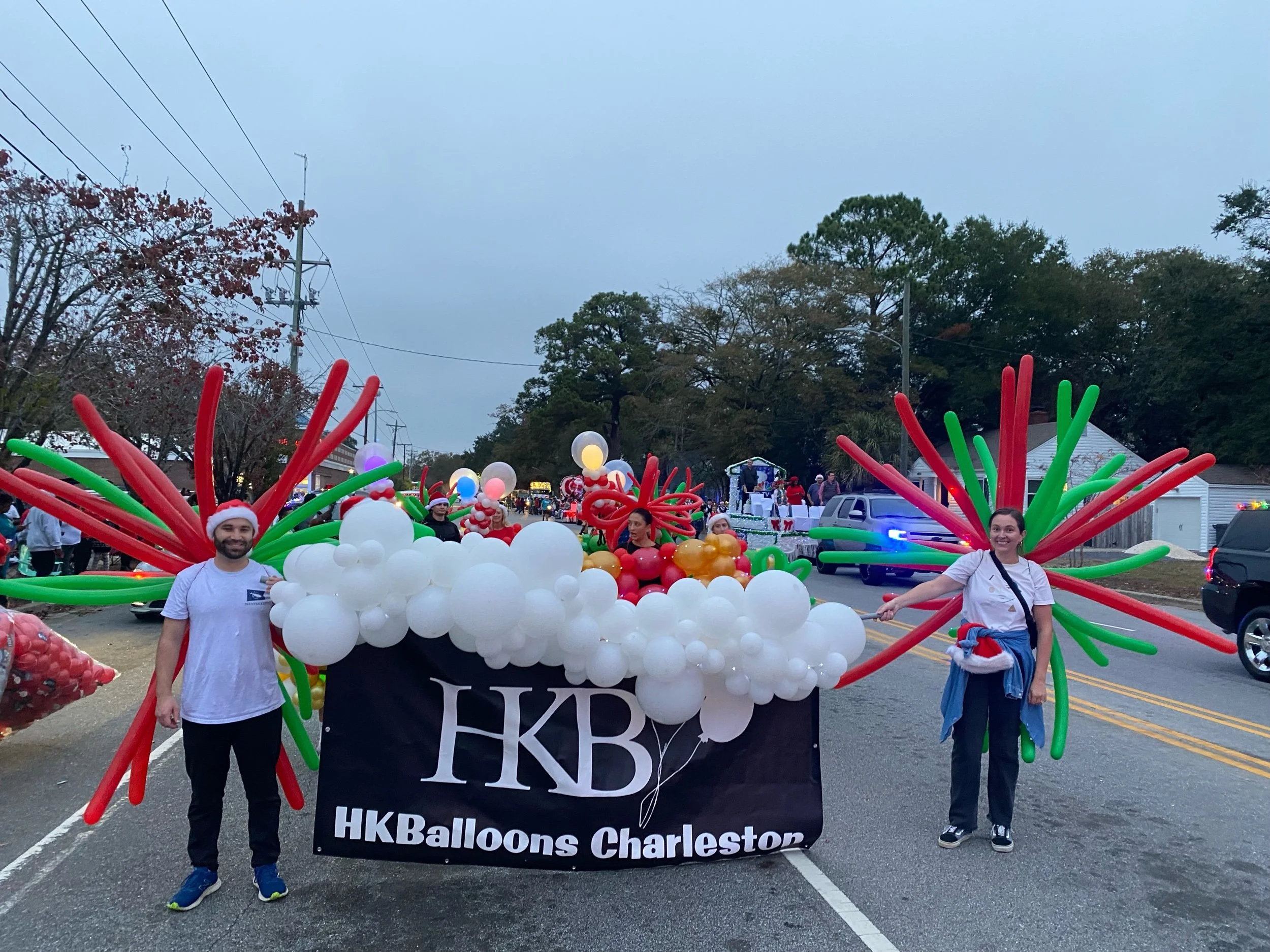 People in a Christmas parade with a float decorated with balloons, labeled 'HKBallons Charleston,' featuring large red and green balloon art resembling poinsettia leaves, and people dressed in holiday attire.