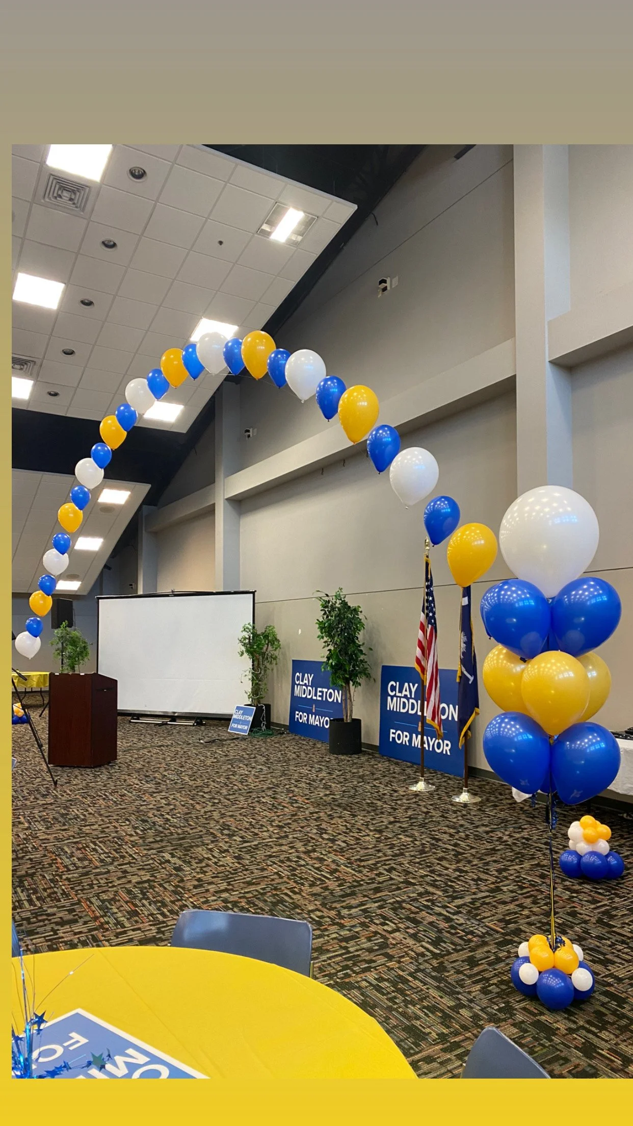 Event setup with blue, white, and yellow balloons arranged in arches and clusters, campaign signs for Clay Middleton, an American flag, and campaign materials at a political gathering or rally.