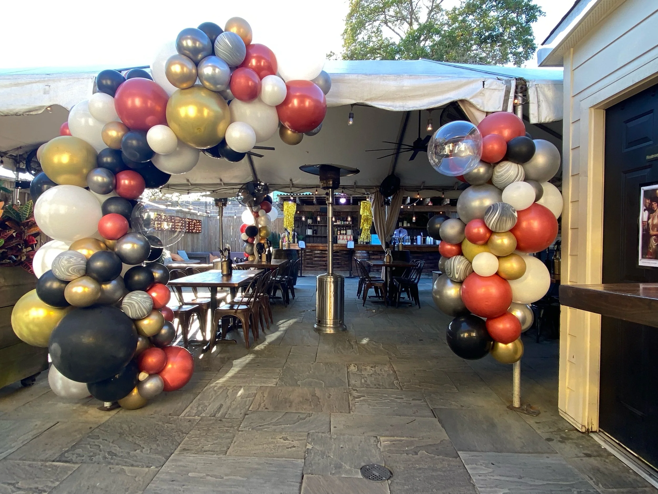 Decorative balloon arch in black, gold, white, and red at restaurant entrance with outdoor seating visible in background.