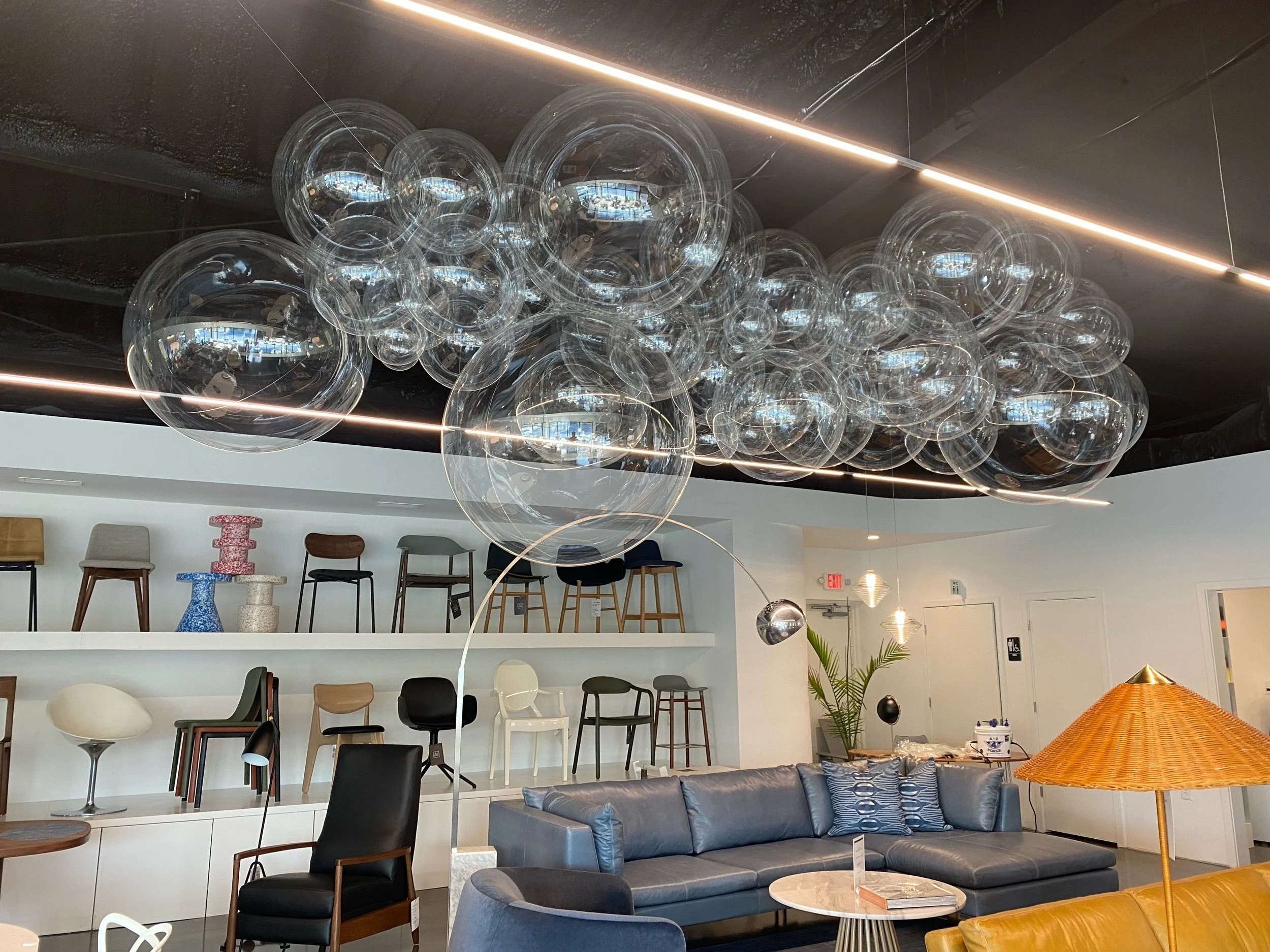 Modern interior with a black ceiling feature, hanging glass bubble chandelier, navy blue sofa, and various chairs on display, with shelves of chairs and a large wicker lamp.