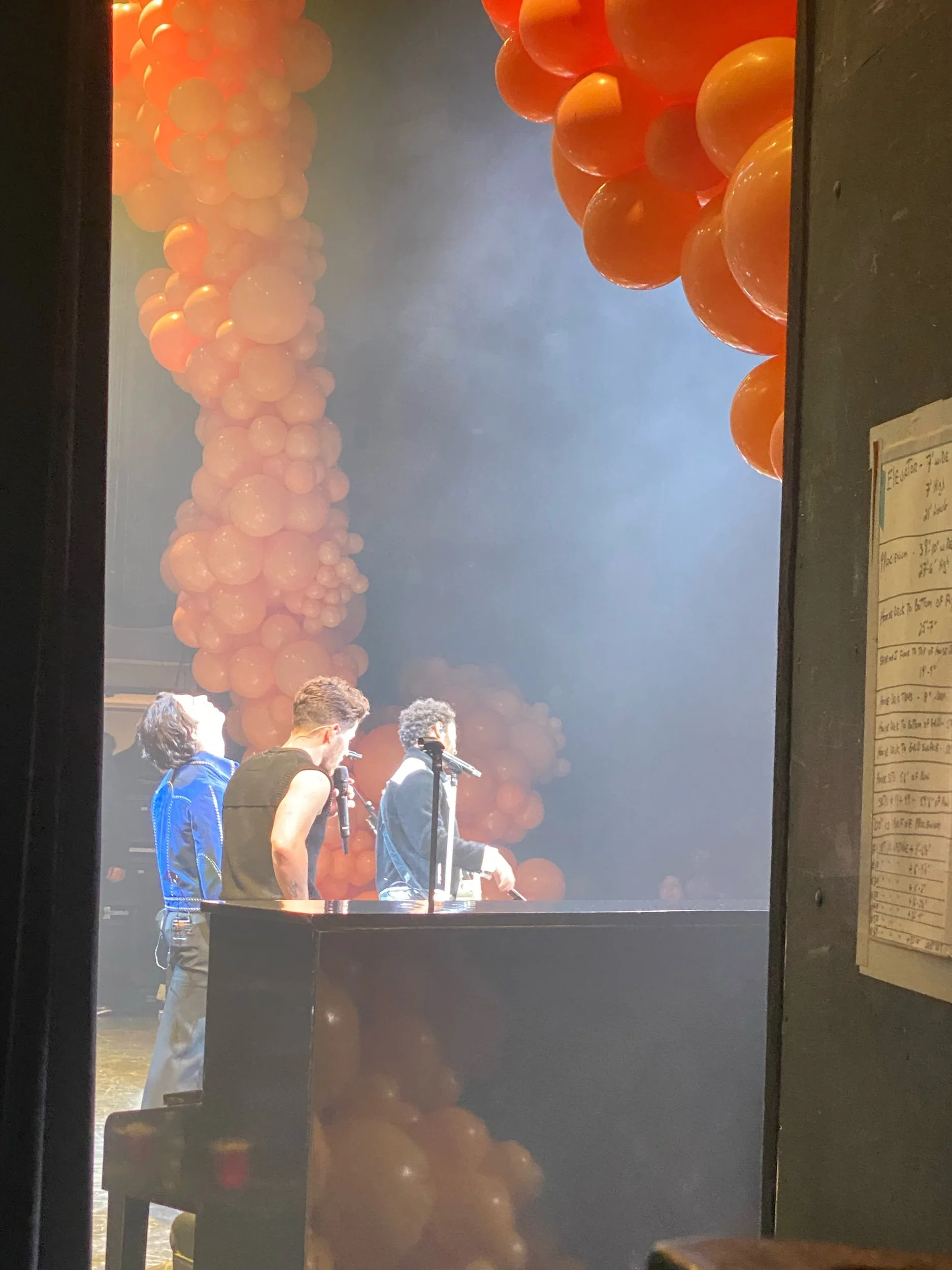 The Jonas Brothers on a stage with orange balloon decorations in the background, seen from behind a door or opening.