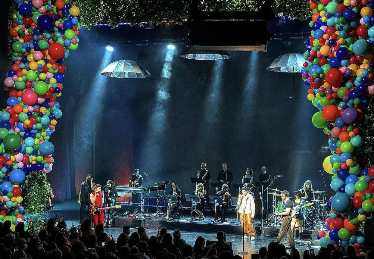 The Jonas Brothers perform at a concert stage decorated with large colorful balloon pillars on both sides, with a musical band performing under blue stage lights and audience members watching.