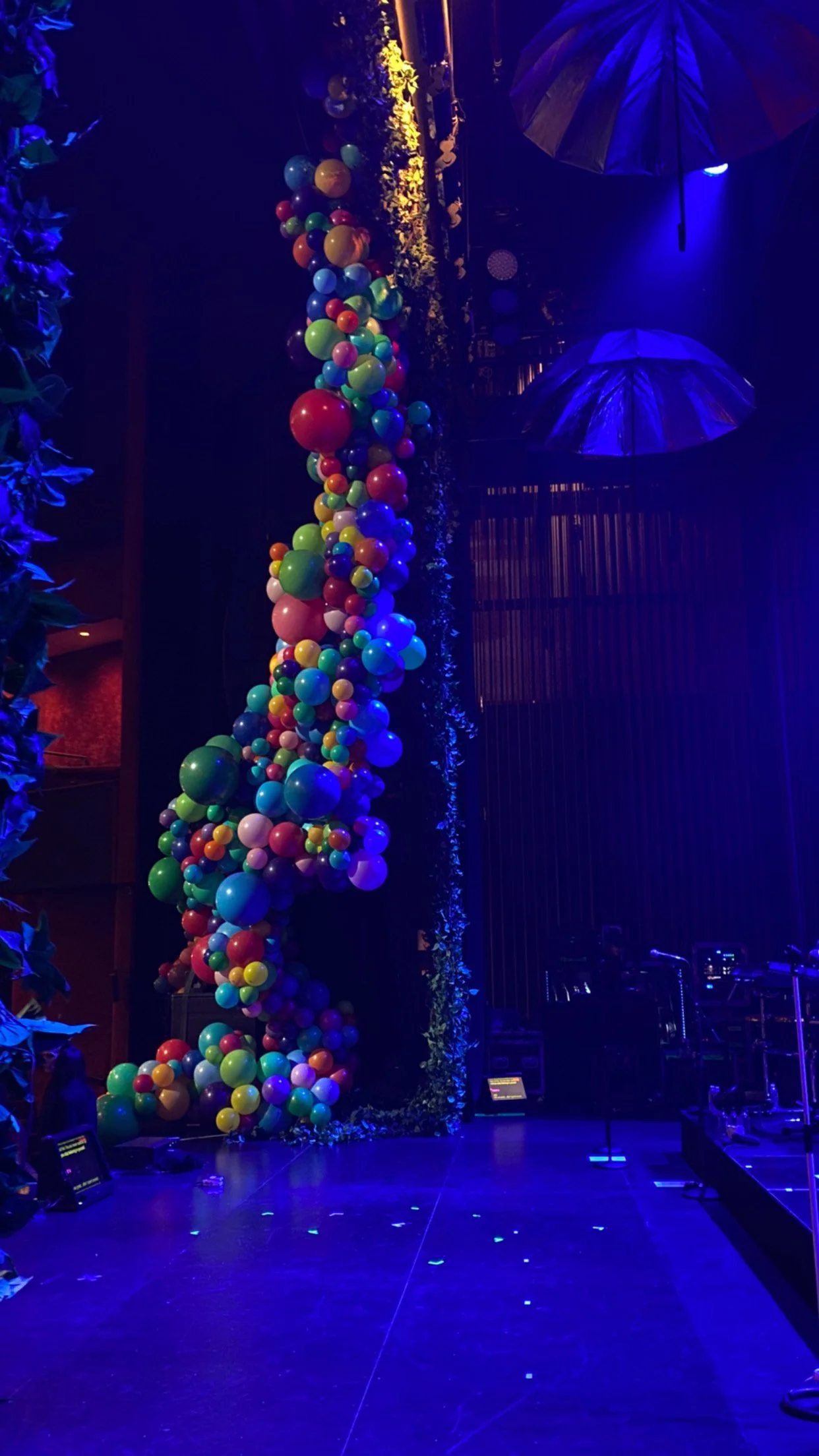 The Jonas Brothers perform on a stage with colorful balloons, including red, green, yellow, blue, purple, & white, arranged vertically on a stage. Large umbrellas hang from the ceiling, and green foliage decorates the edges.