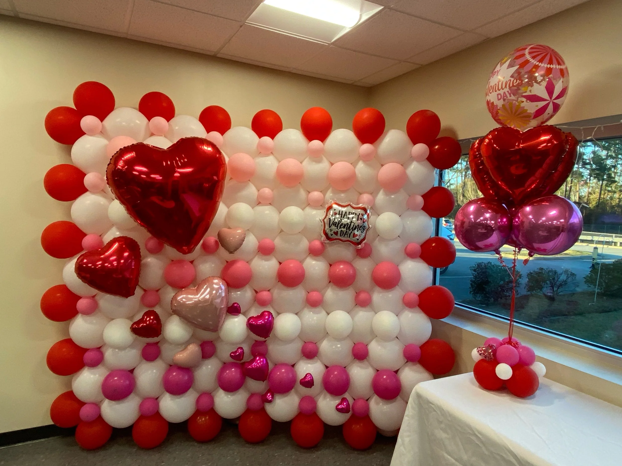 Decorative backdrop with white, pink, and red balloons arranged for Valentine's Day, including several heart-shaped balloons and a sign that says "Happy Valentine's Day." Pink, red, and purple balloons are also on a table near a window.