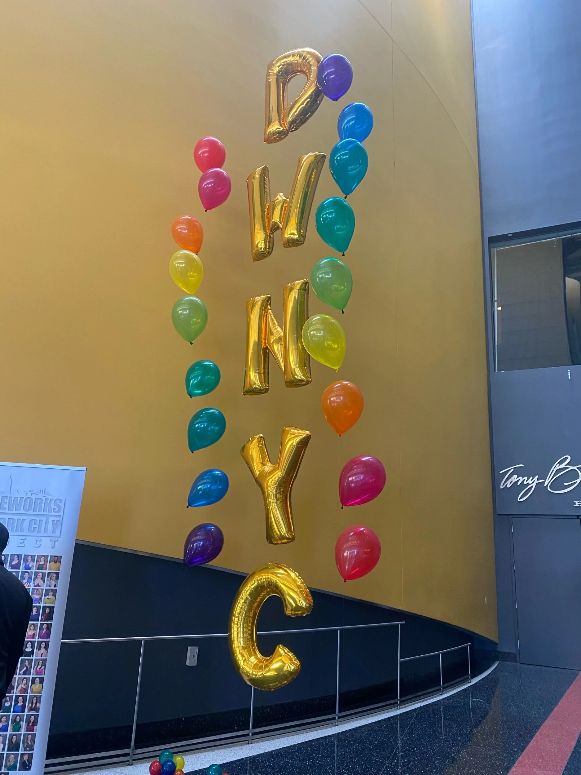 Colorful balloons arranged with large gold-letter balloons spelling 'D W W K Y' hanging against a gold wall.