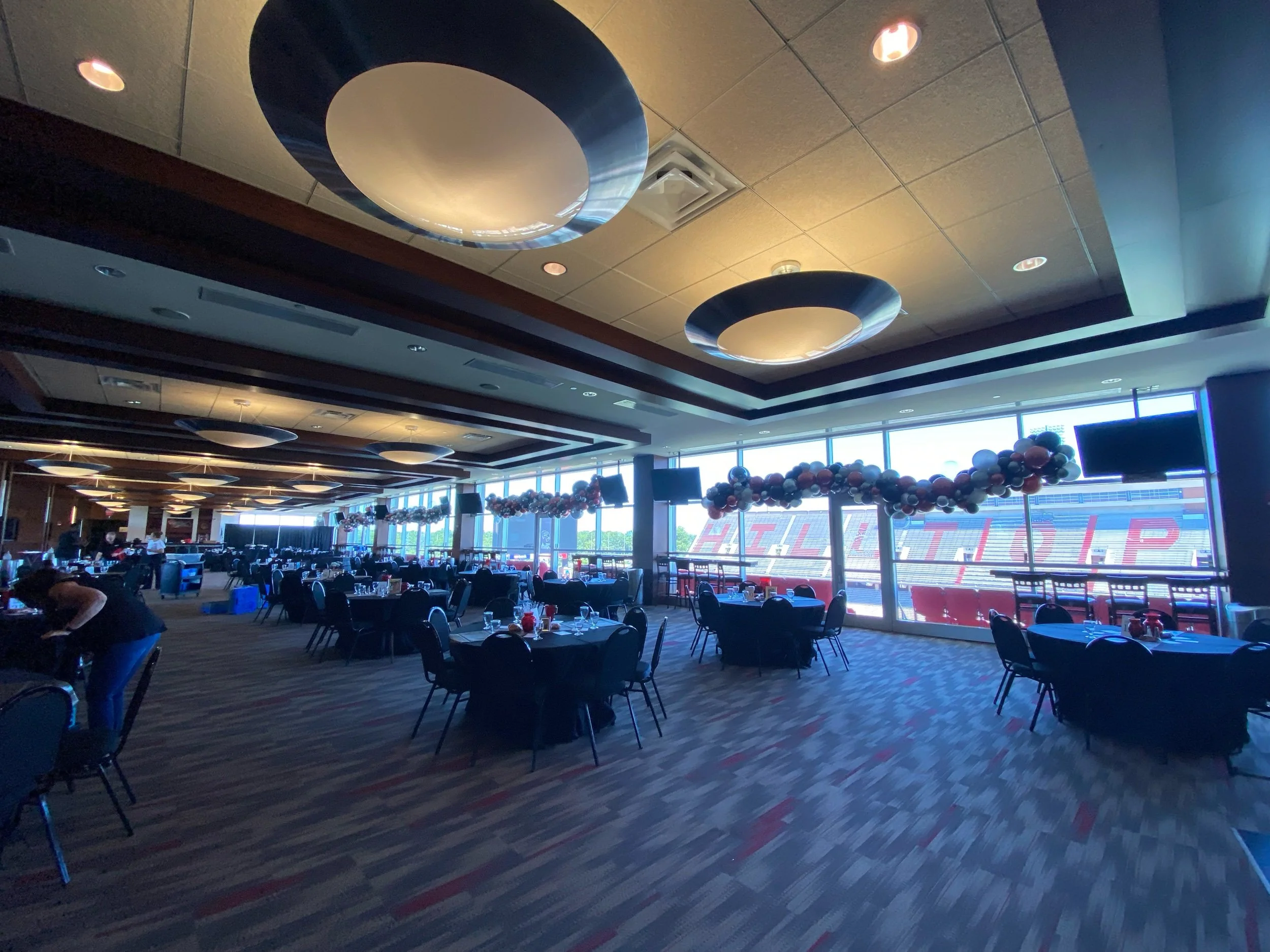 Large indoor event space decorated with balloons and set with round tables and chairs, with stadium seating visible through large windows.