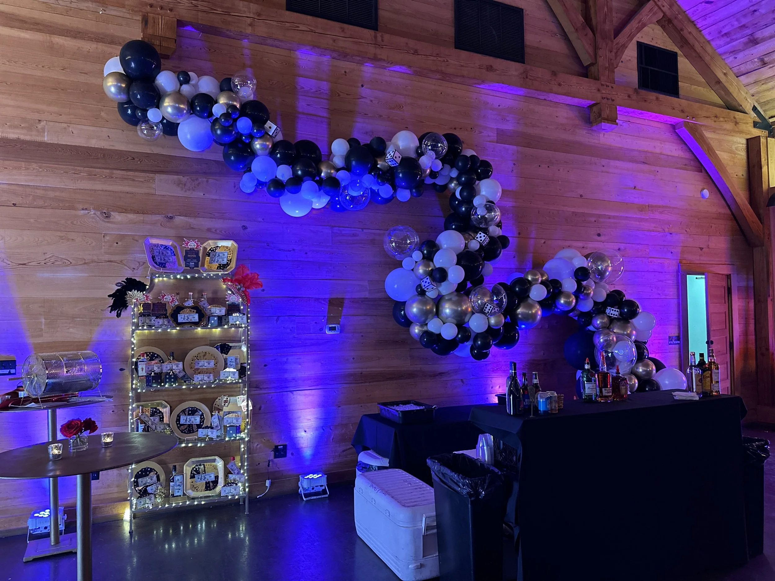 Decorative balloon arch with black, white, gold, and clear balloons, along with dice and star-shaped decorations, set against a wood-paneled wall. A bar with bottles and a shelving unit with treats and signs are also visible in a dimly lit room with