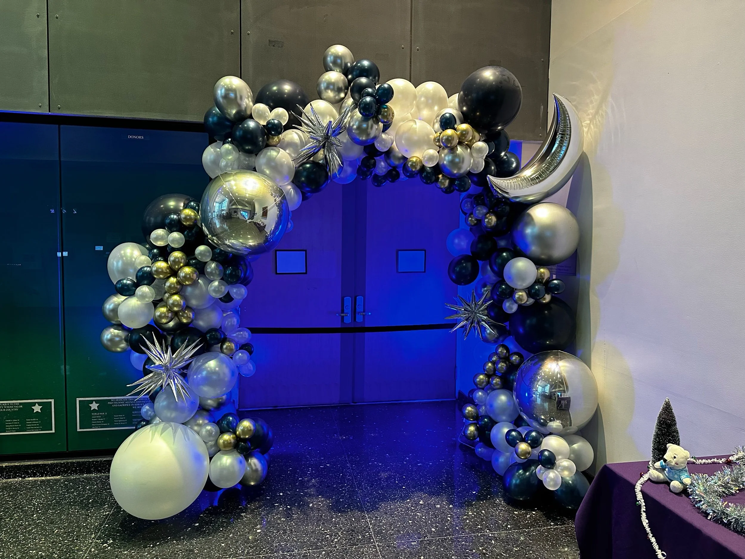 Decorative balloon arch with black, white, gold, and silver balloons, including star-shaped and crescent moon-shaped balloons, at an indoor event entrance.