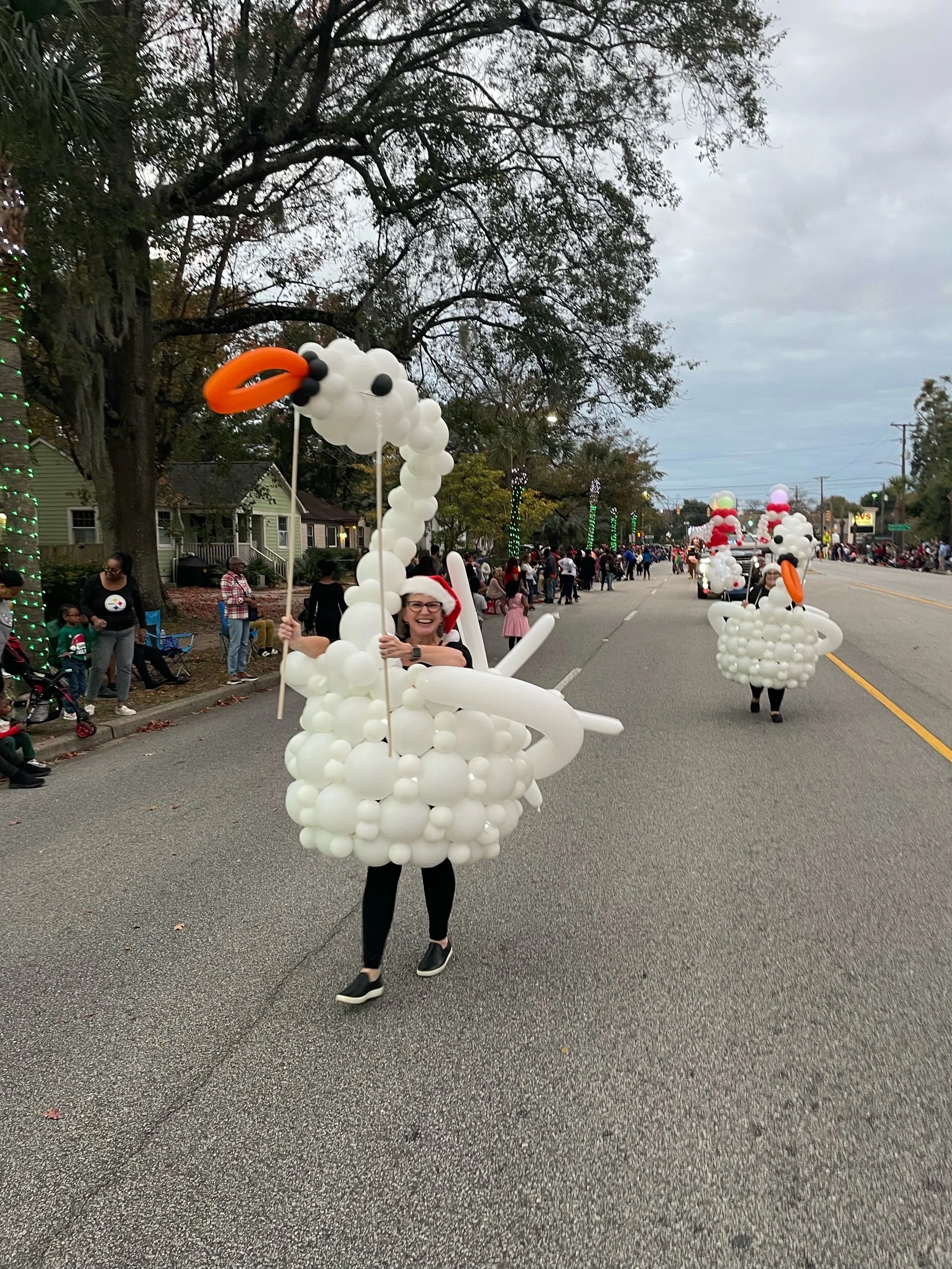 Person dressed in a swan costume made of balloons, participating in a parade, with other balloon swan costumes and spectators along the street.