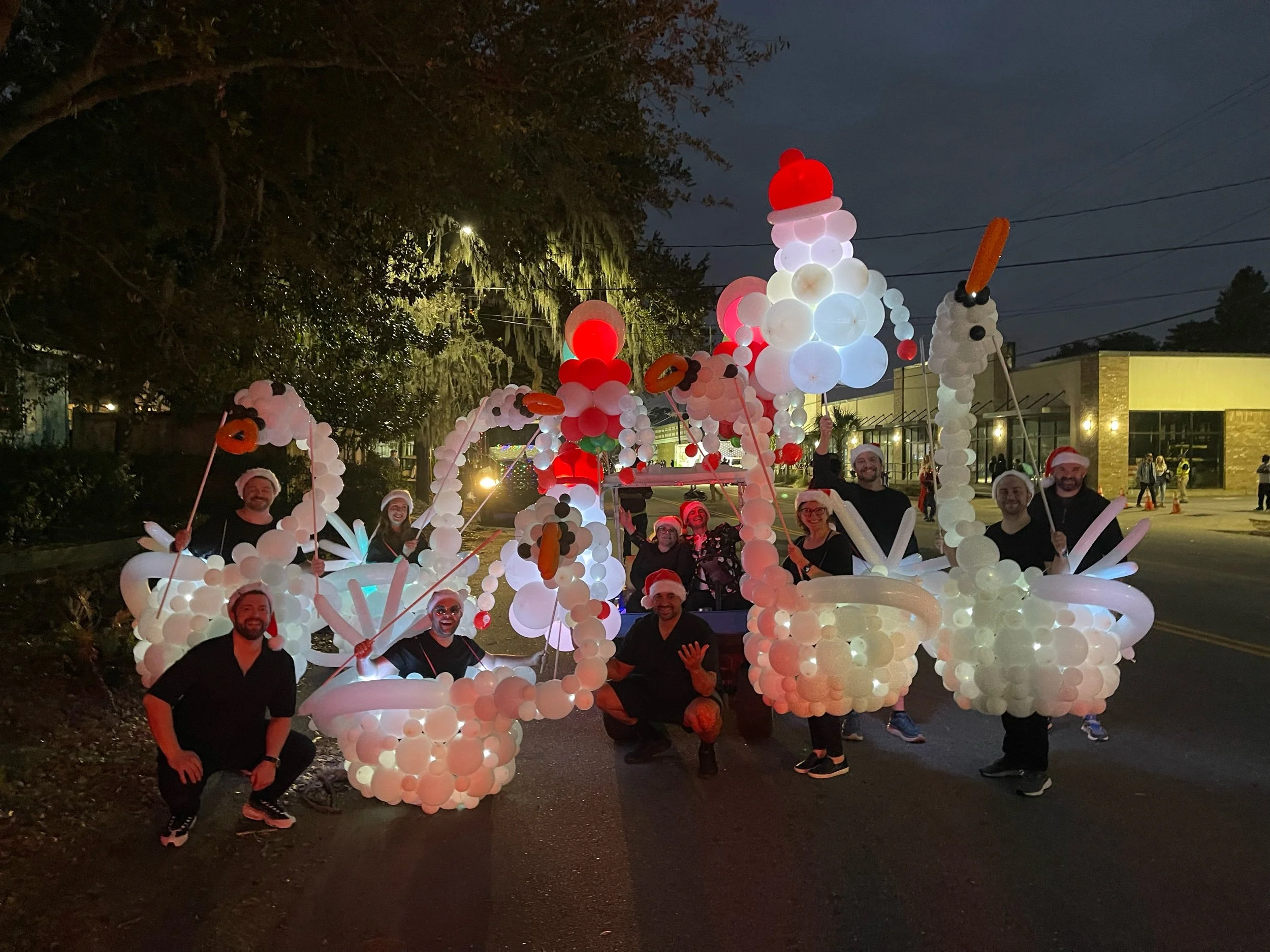 Group of people dressed in black wearing Santa hats and smiling, gathered around a festive float decorated with white balloons shaped like swans, geese, and other birds, illuminated at night for a holiday parade.