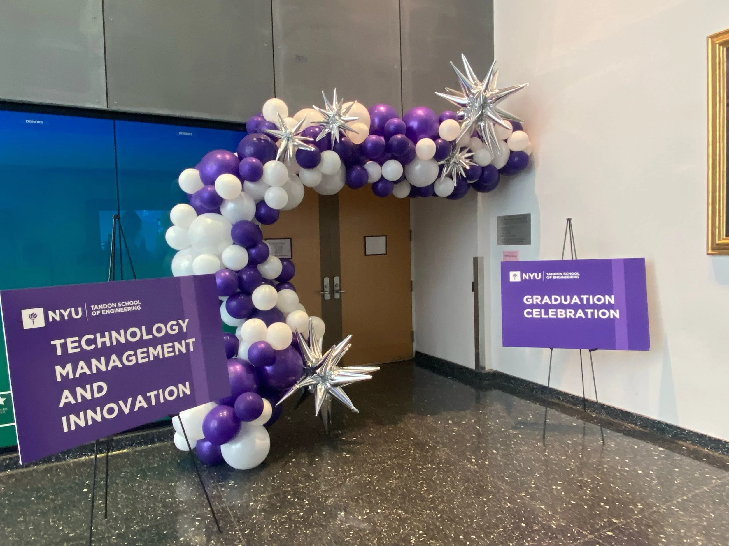 Balloon arch decorated with white, purple, and silver balloons, with signs for NYU Tandon School of Engineering's Technology Management and Innovation graduation celebration.