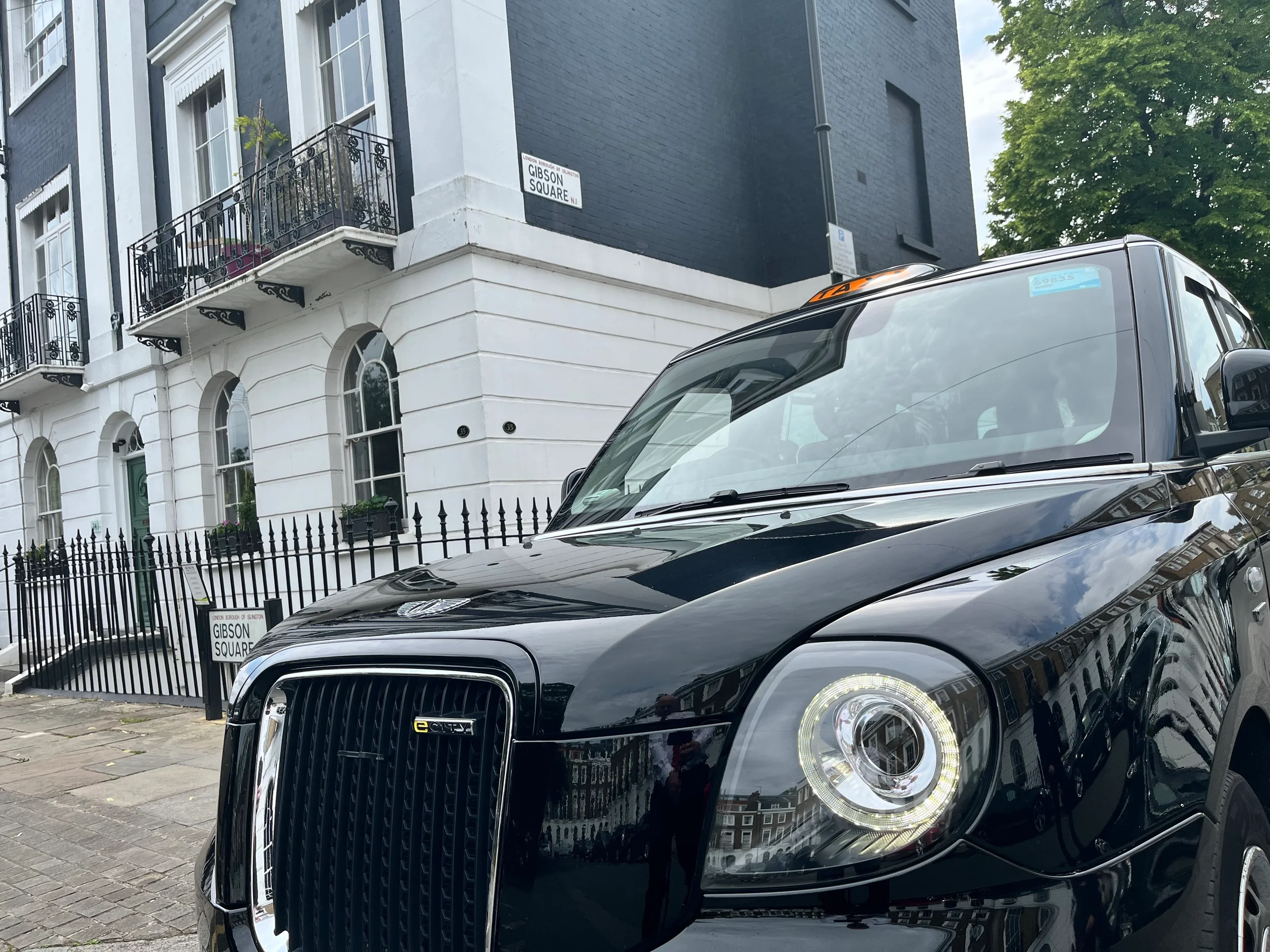 Black taxi parked on the street in front of a white building with balconies and black iron railings, in an urban neighborhood, with a tree in the background.