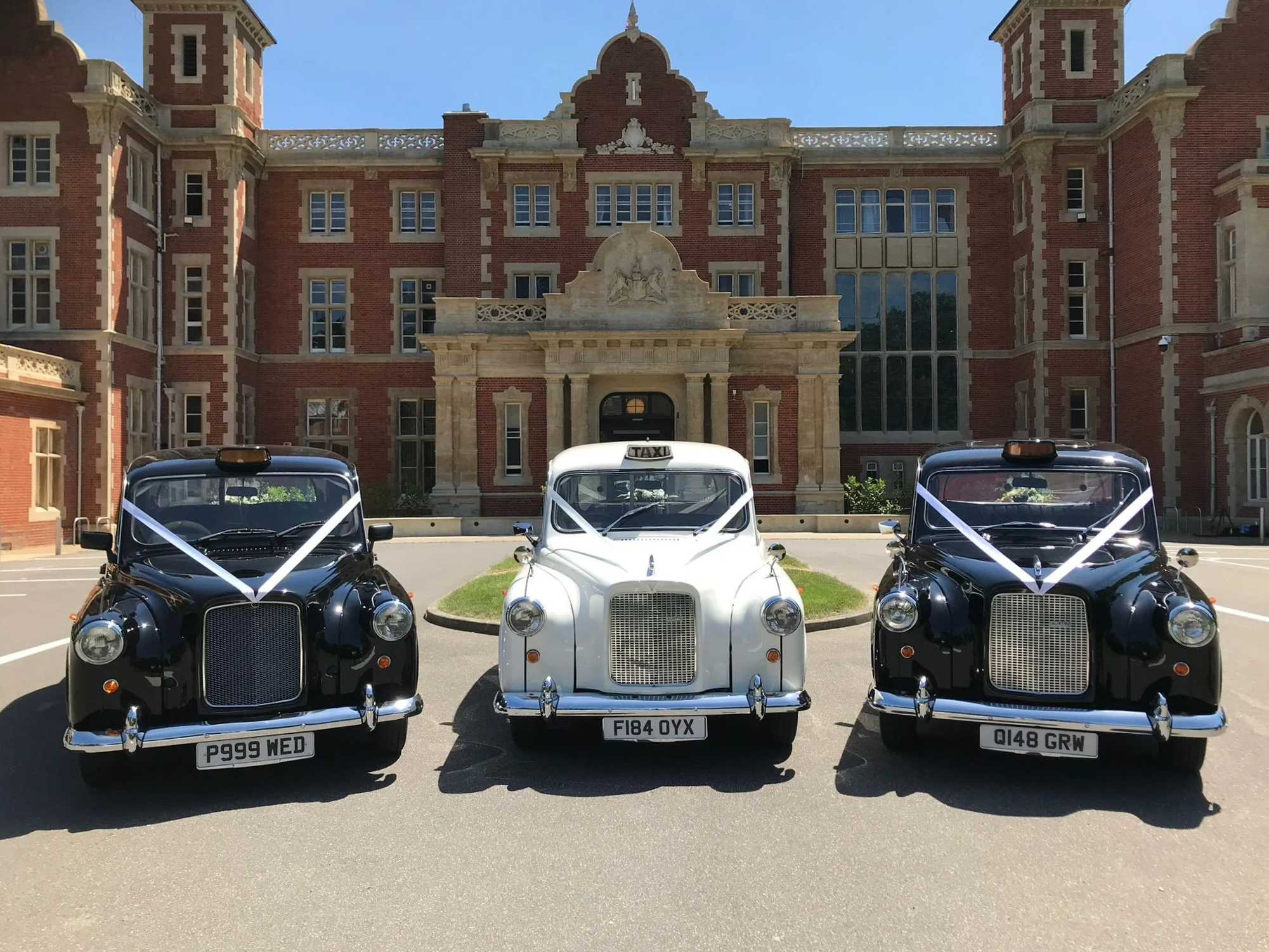 Three vintage wedding taxis parked in front of a large brick and stone historic building, decorated with white ribbons for a wedding.
