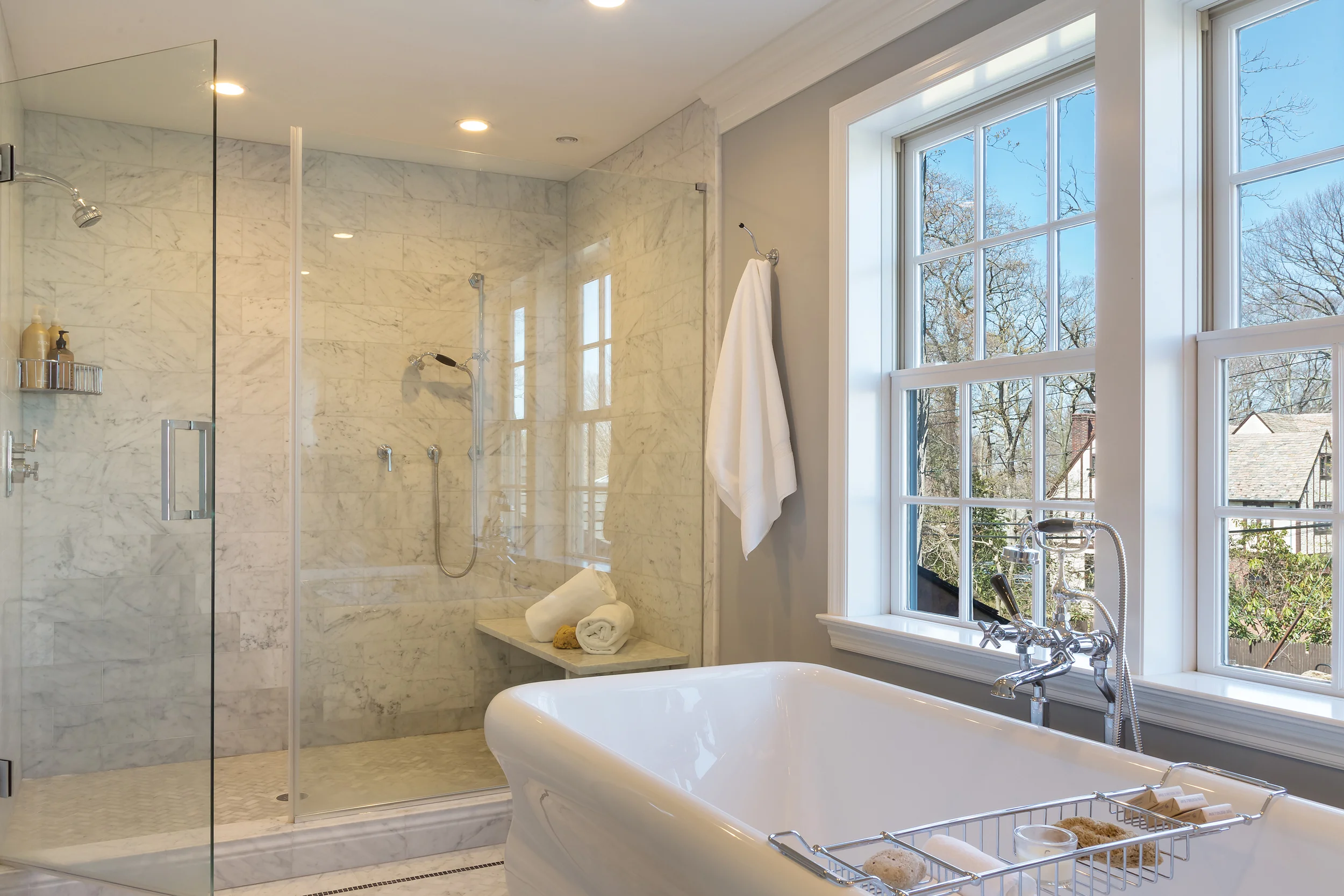Tom Fox designed a spacious shower that runs the width of the room as he did with the vanity wall. Custom-designed Carrara marble throughout with a seamless glass enclosure creates optimal visual space. At one end is a showerhead; at the other, a hand-held shower is flexible whether standing or sitting.