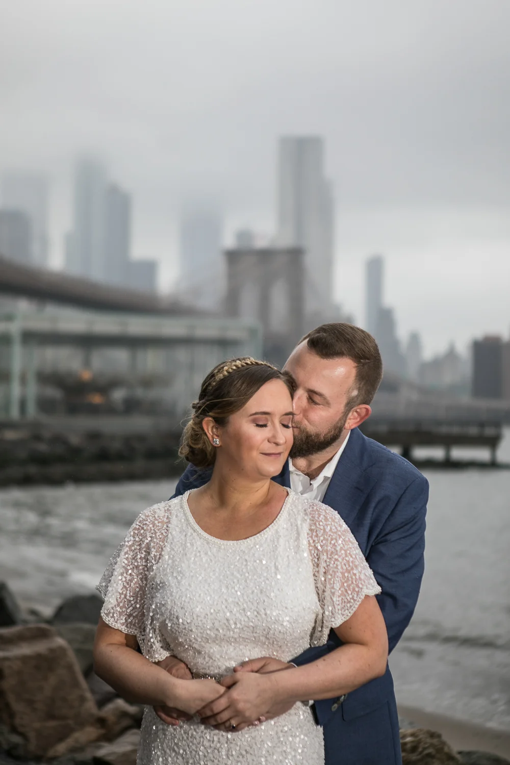 Wedding photos in front of the Brooklyn Bridge. New York Bride in a short wedding dress and groom in a blue suit pose in front of the Brooklyn Bridge. | Intimate Wedding at the Malthouse in Manhattan, New York City.