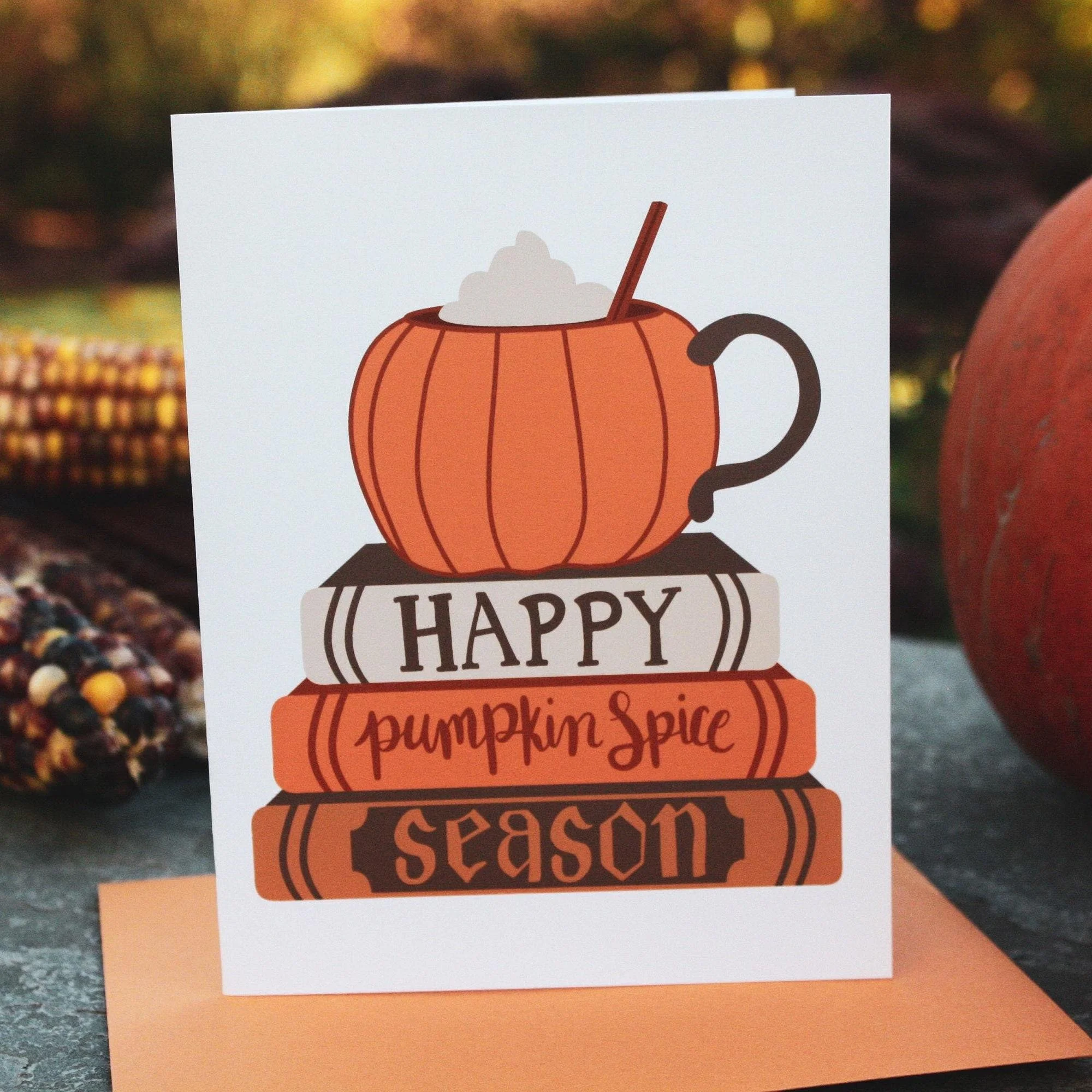 Greeting card with a pumpkin shaped mug atop a stack of books with the message "Happy Pumpkin Spice Season" on the book spines