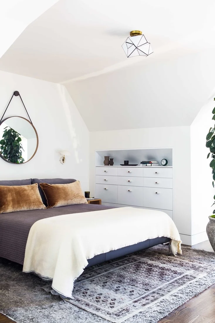 How to decorate around a bed. Option: Hang a mirror above the bed. (Design: Lauren Geremia via Architectural Digest) // gray bedroom, round mirror in bedroom, captain's mirror in bedroom, angled ceiling bedroom