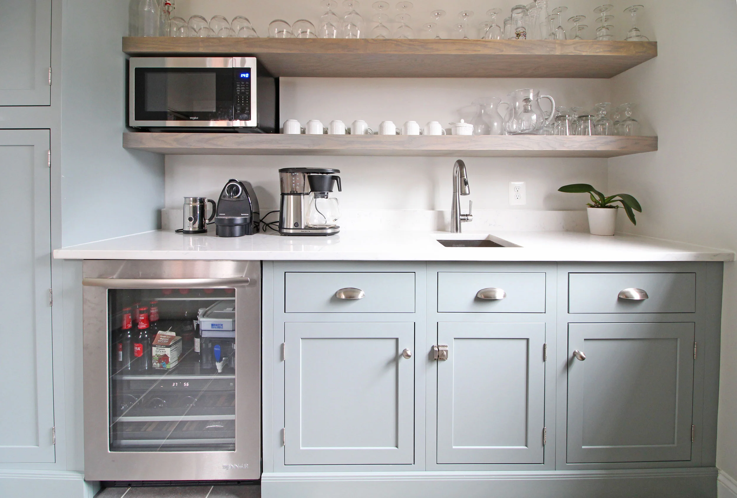 Light, blue-green cabinets in this transitional farmhouse butler's pantry perfectly complement the blue glass subway tile in the neighboring kitchen. Open shelving provides great storage without the weight of upper cabinets, and a beverage fridge and small sink add functionality. 