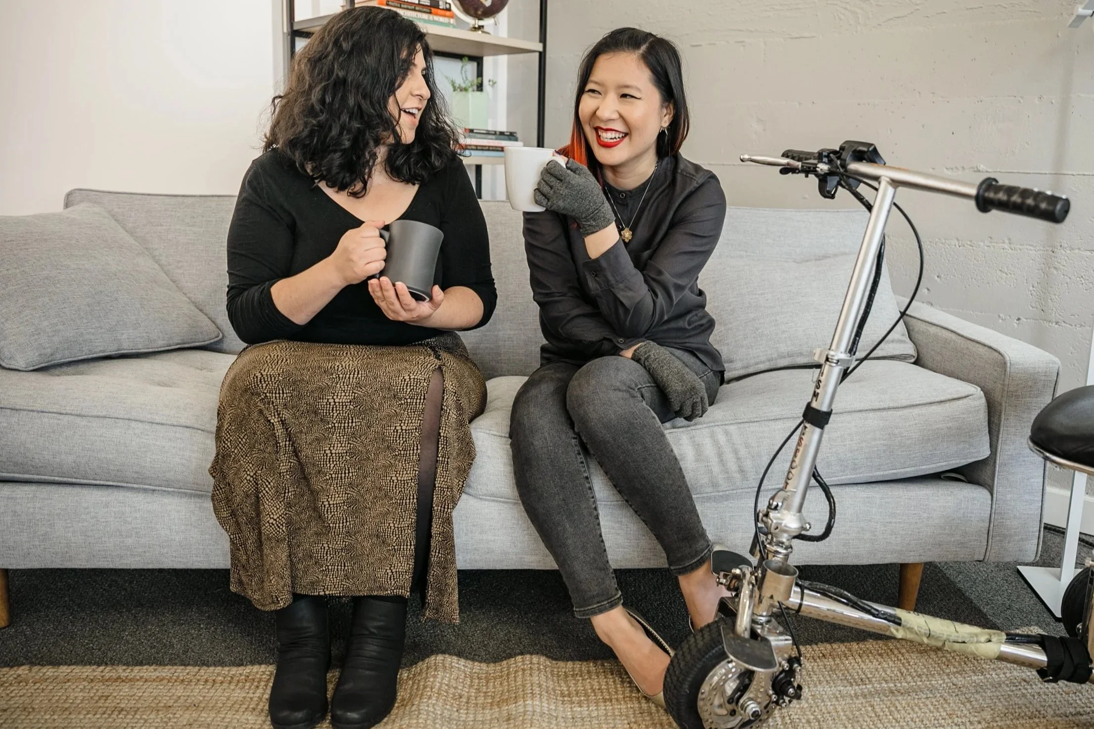 A Latinx disabled woman and an Asian disabled genderfluid person chat and sit on a couch, both holding coffee mugs. An electric lightweight mobility scooter rests on the side. Photo provided by the Disabled and Here project.