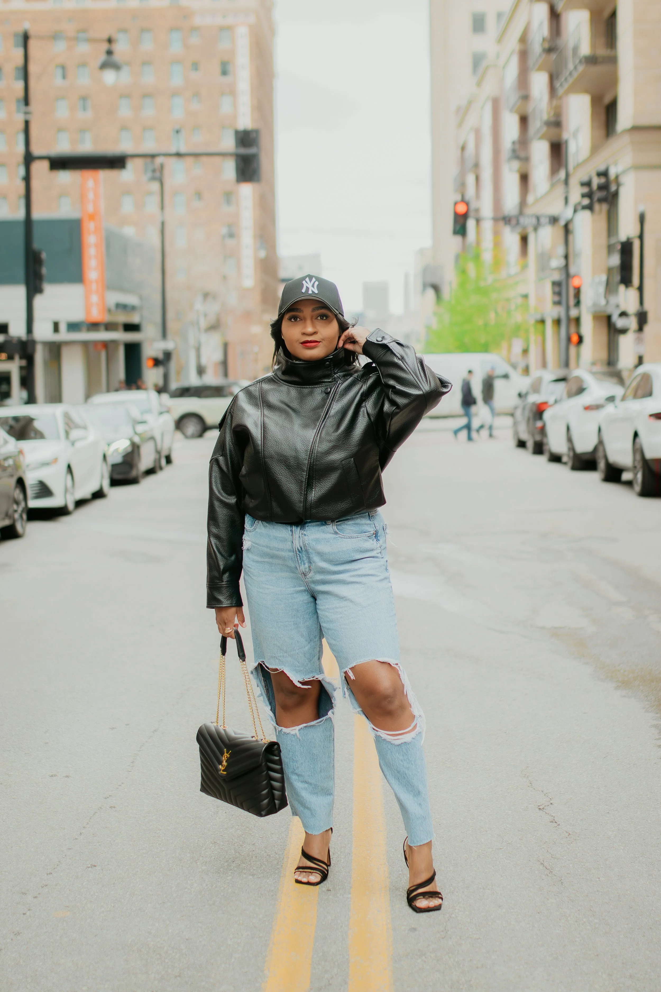 Woman wearing a black tank top, oversized black leather jacket, ripped jeans, NY fitted cap, and black strappy heels for a chic street style outfit. in kansas city