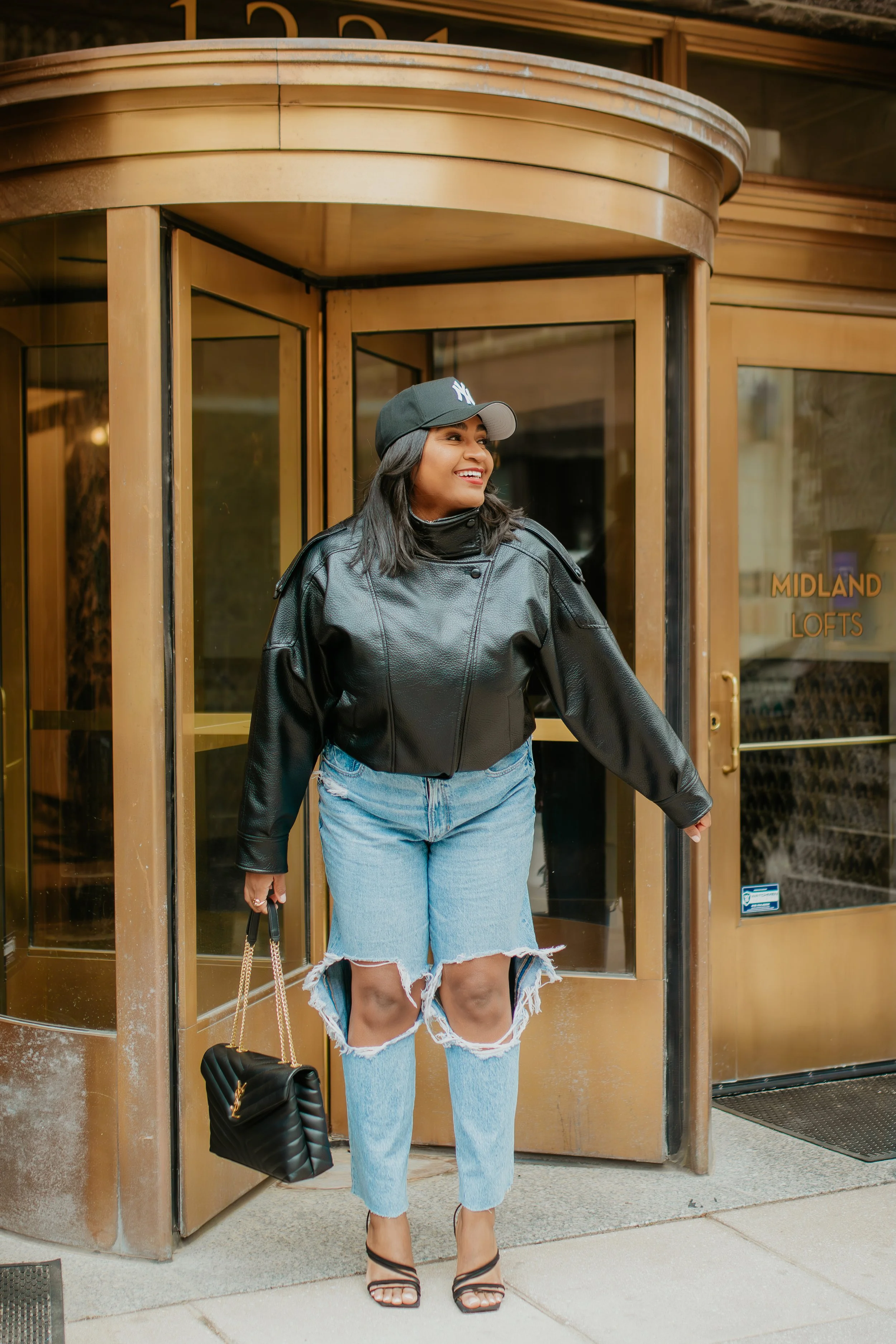 Woman wearing a black tank top, oversized black leather jacket, ripped jeans, NY fitted cap, and black strappy heels for a chic street style outfit.