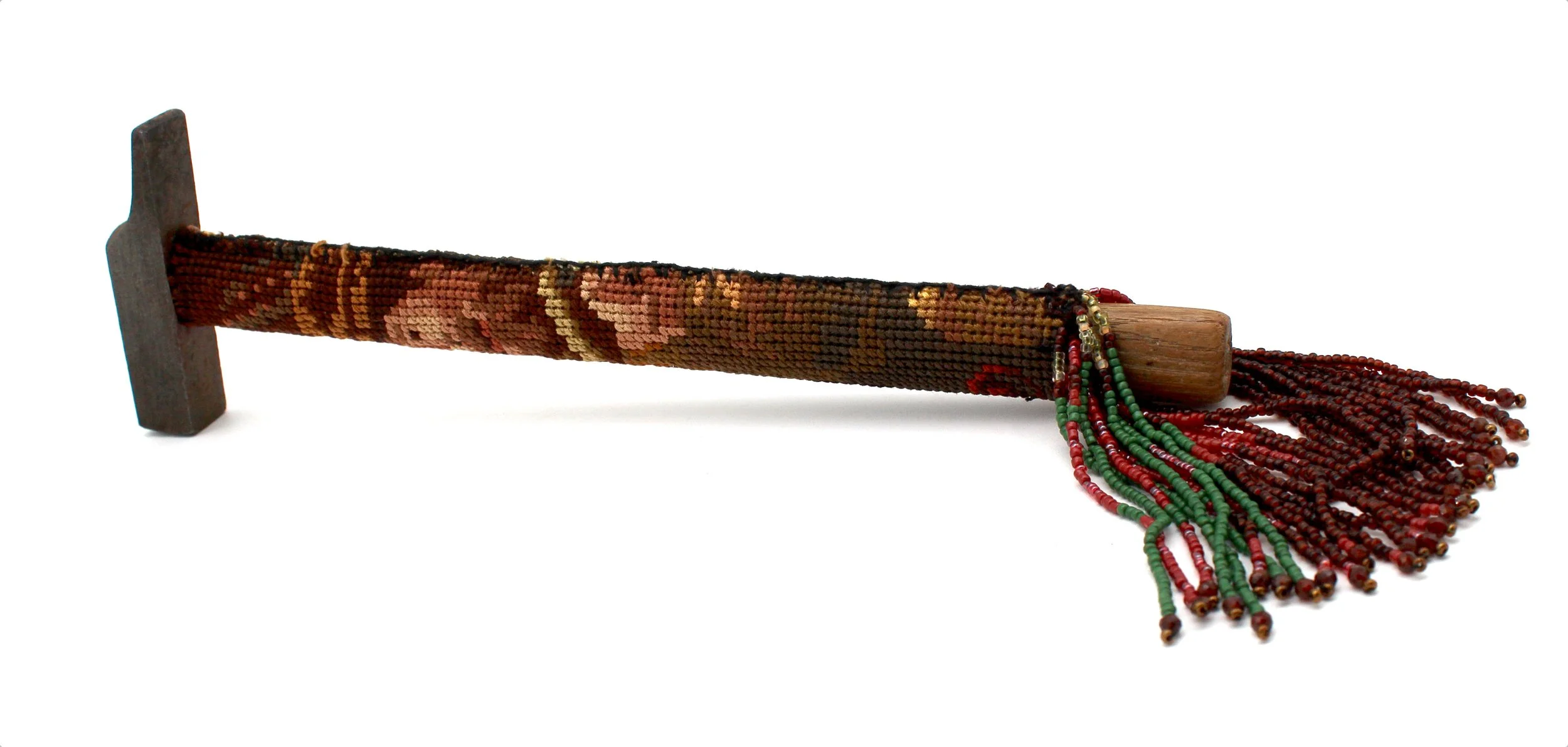 A cross peen hammer sitting on its side. The handle is covered in an earth tone cross-stitch and there is a four inch bead fringe in purple, green, and brown hanging off the end.