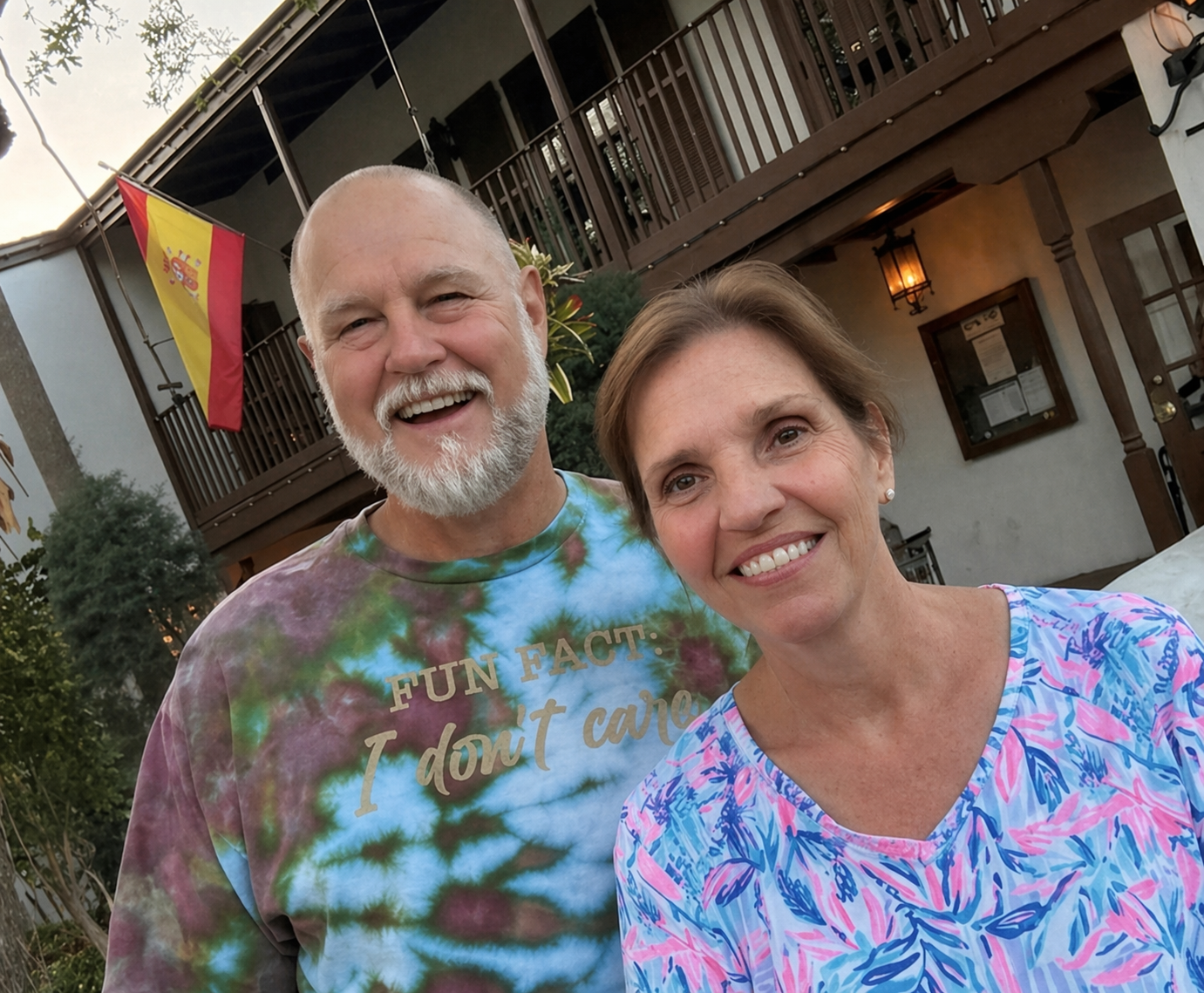 A smiling older man with a beard and a tie-dye shirt standing next to a smiling woman with short hair and a blue patterned shirt, outdoors in front of a building with a Spanish flag hanging from the balcony.