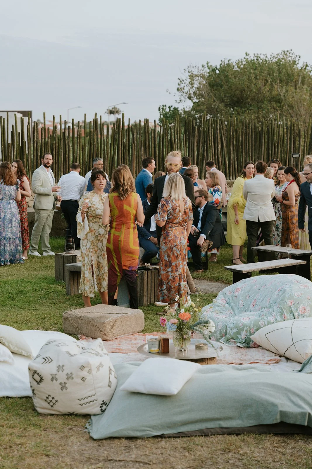 People gathered outdoors at a social event, with sofas, pillows, and flowers in the foreground and a wooden fence and trees in the background.