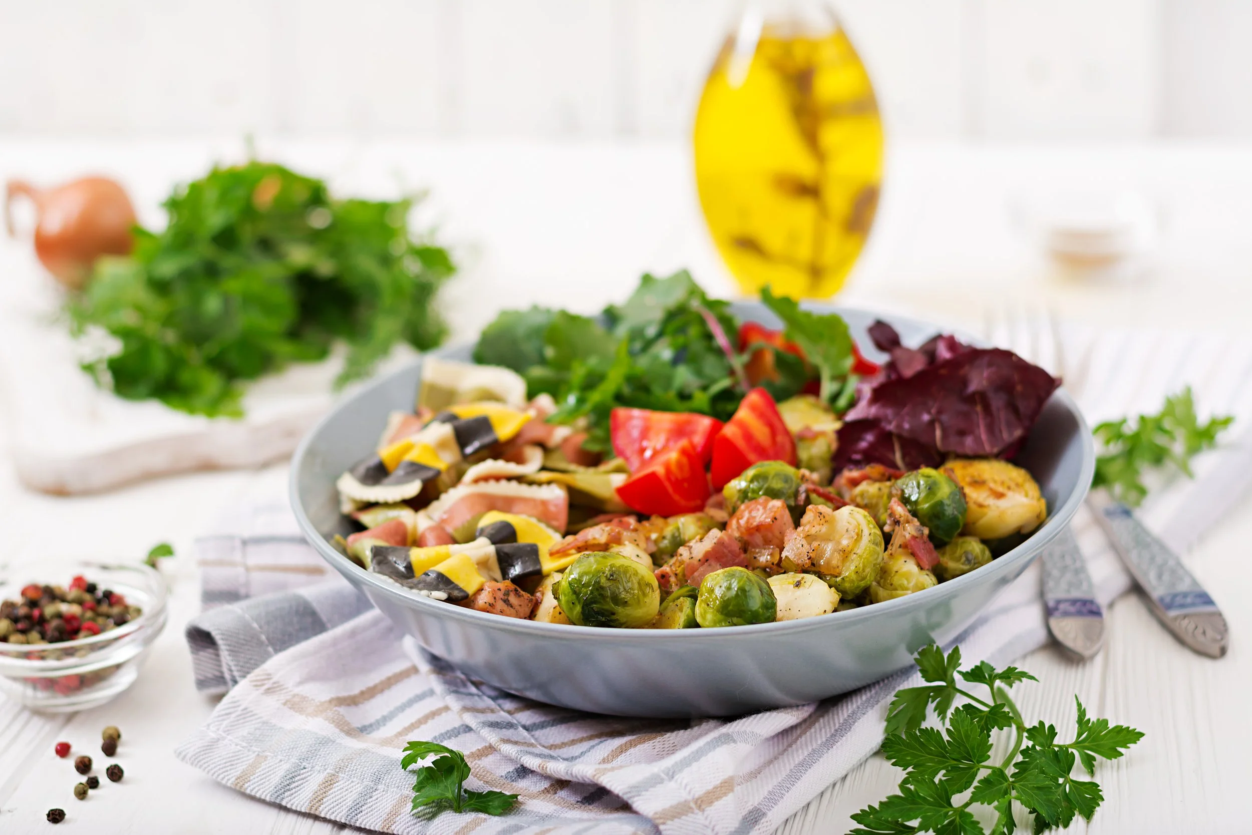 A large bowl of mixed salad with tomatoes, Brussels sprouts, lettuce, and other vegetables on a white wooden table, with a bottle of olive oil, a fork, a knife, and some parsley nearby.