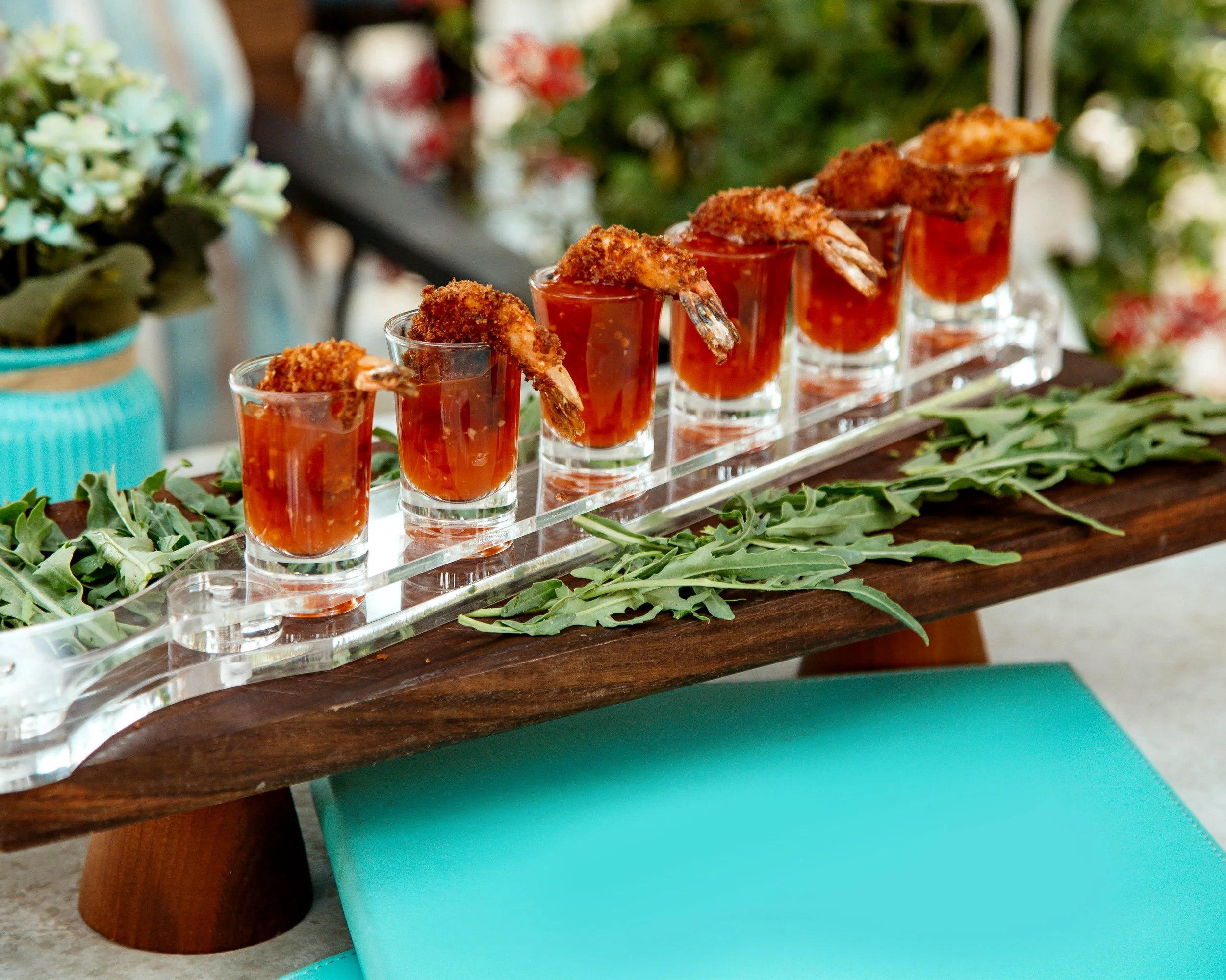 Five shrimp cocktails served in small glasses with crushed crackers on top, arranged on a glass tray on a wooden board, decorated with green leafy garnish, with a background of flowers and greenery.