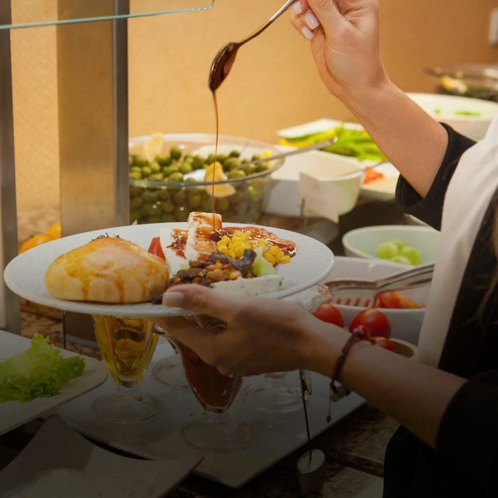 Person pouring chocolate sauce over a dessert on a white plate at a buffet table.