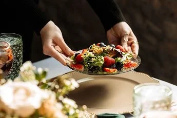 Person serving a plate of salad with tomatoes, lettuce, and other vegetables at a table with flowers and drinks.