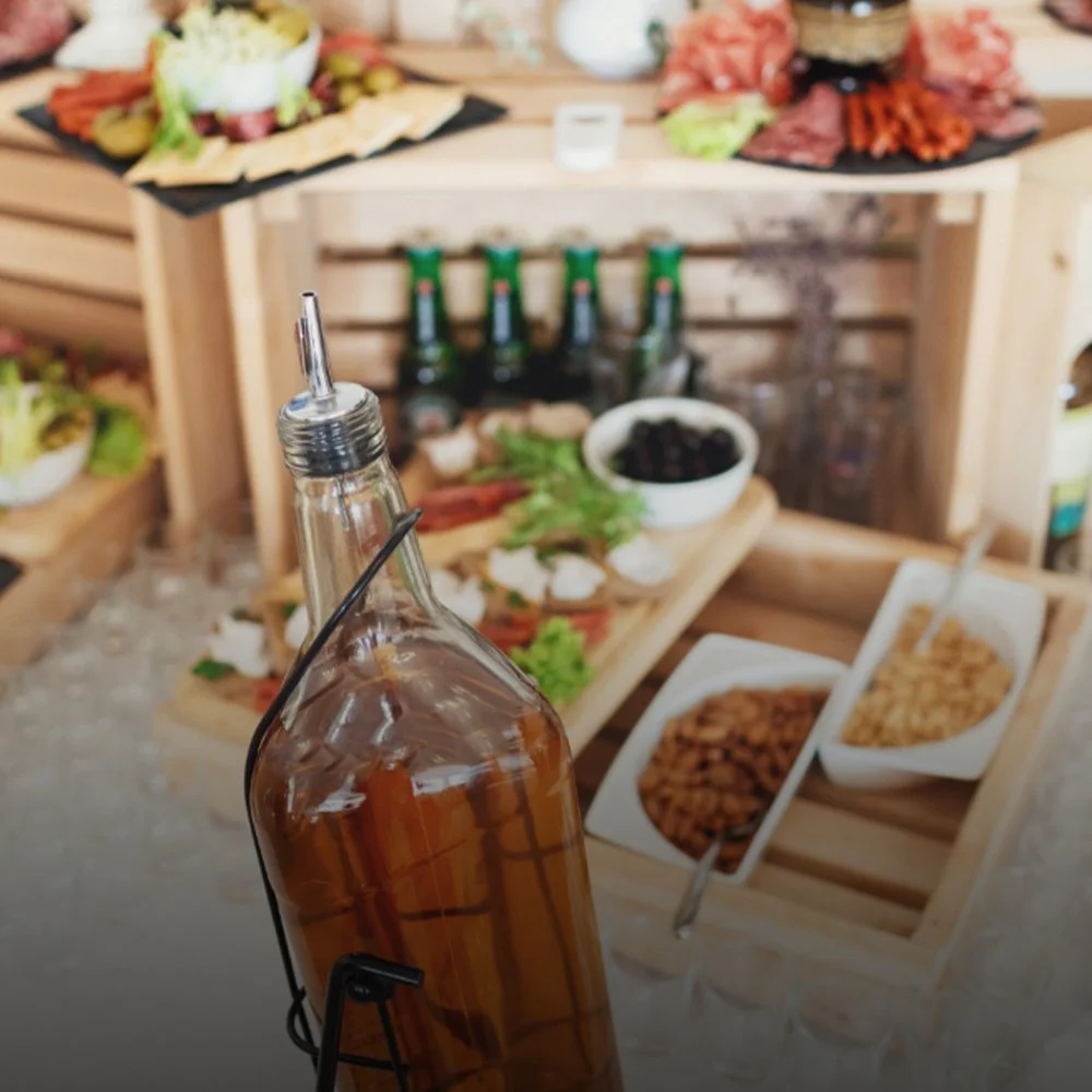 Bottle of brown beverage in foreground with a spread of snacks, dips, and condiments on wooden displays in the background.
