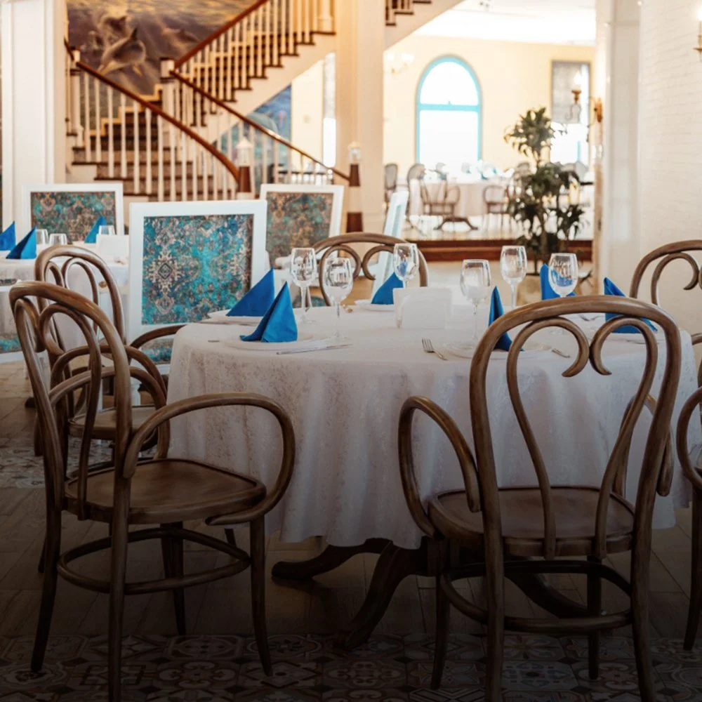 Elegant dining room with round table set for a formal meal, including blue napkins, wine glasses, and white plates, with wooden chairs and a staircase in the background.